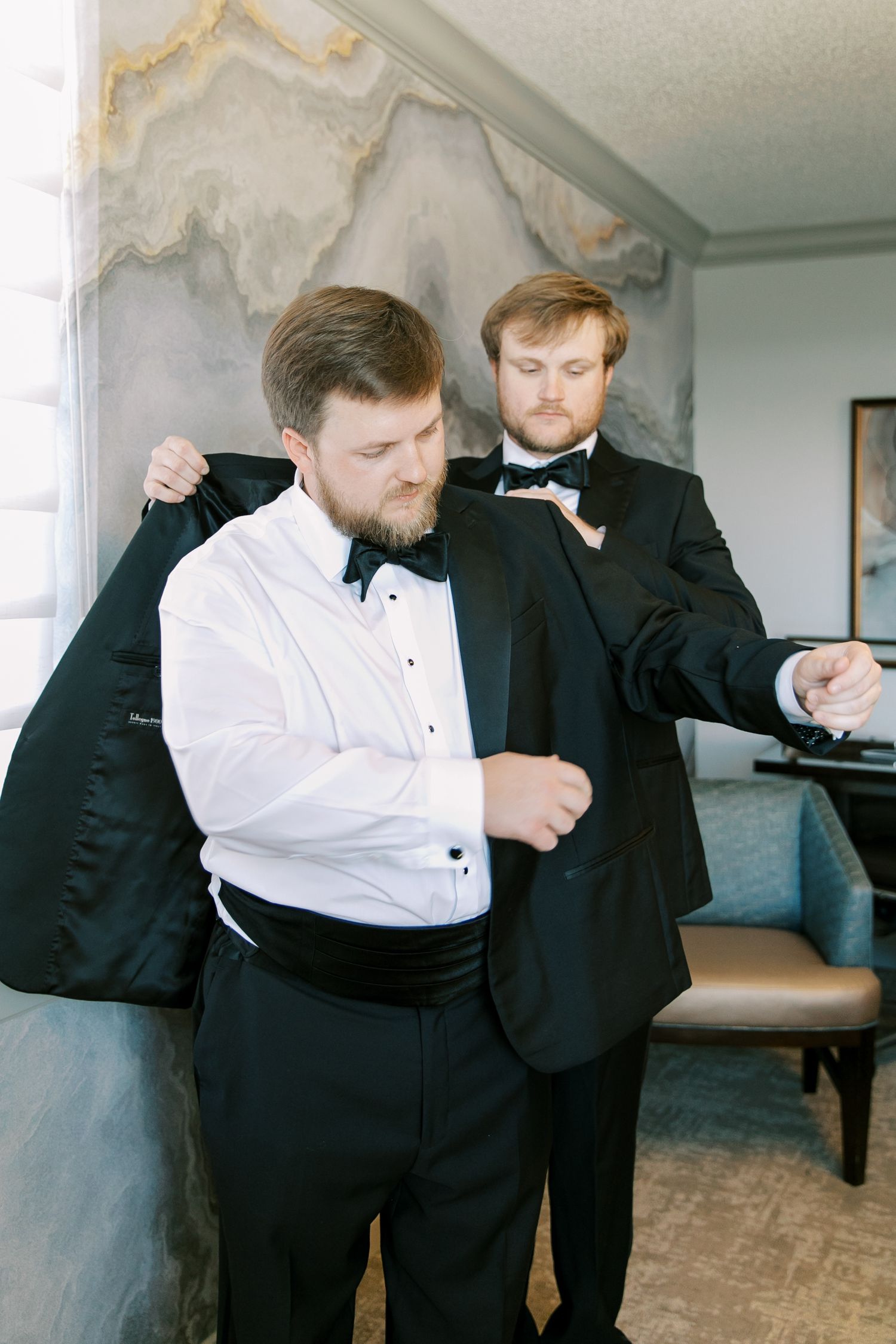 Two men in formal black and white tuxedos getting dressed and helping each other prepare for a formal event.