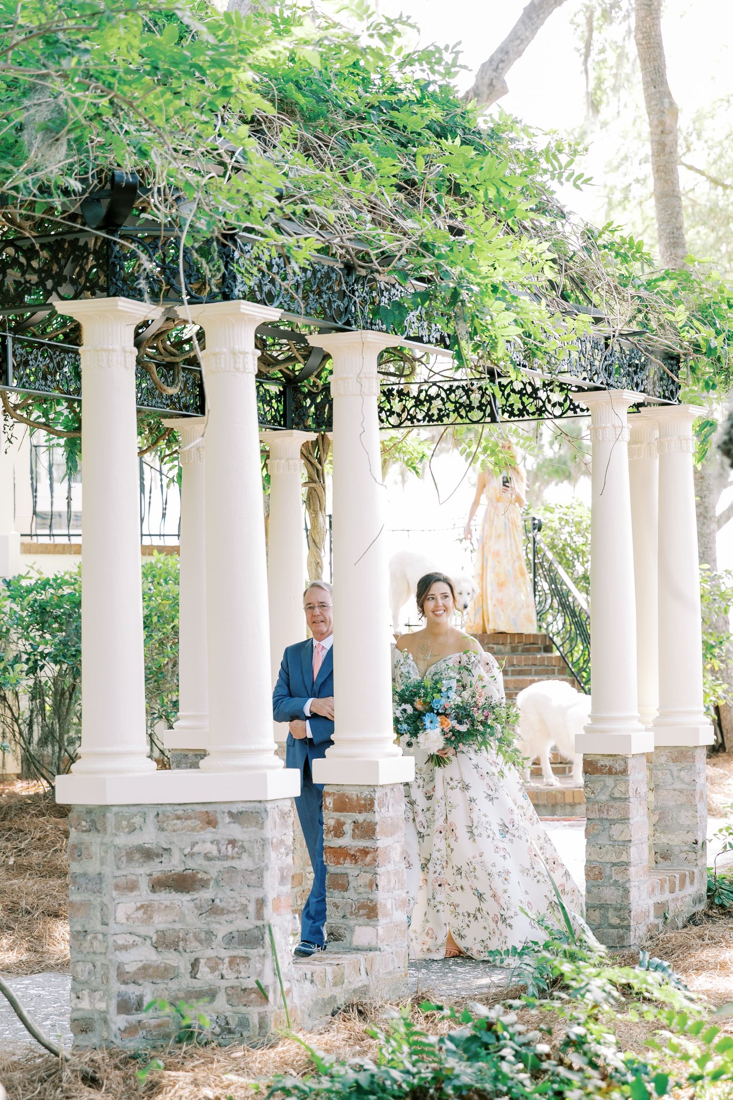 A wedding couple poses under white columns and greenery in an elegant garden setting.