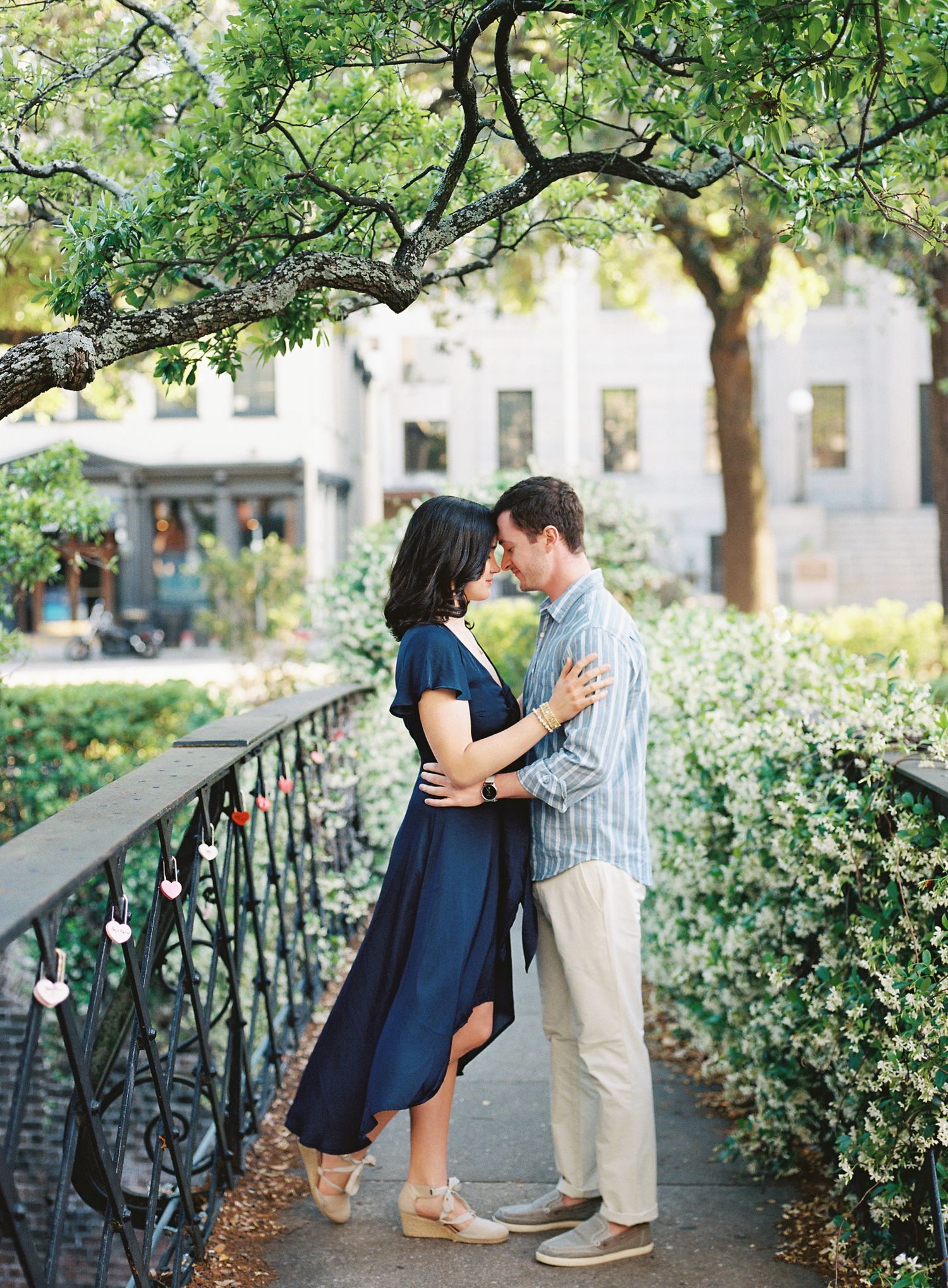 A romantic couple embraces on a garden path under overhanging trees and wrought iron railings.