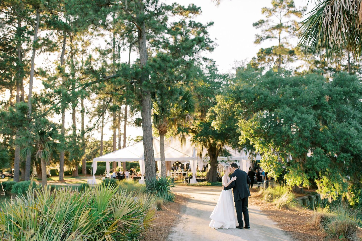 Wedding couple walking along palm-lined path at sunset with reception tent in background at Oldfield golf Club in Bluffton.