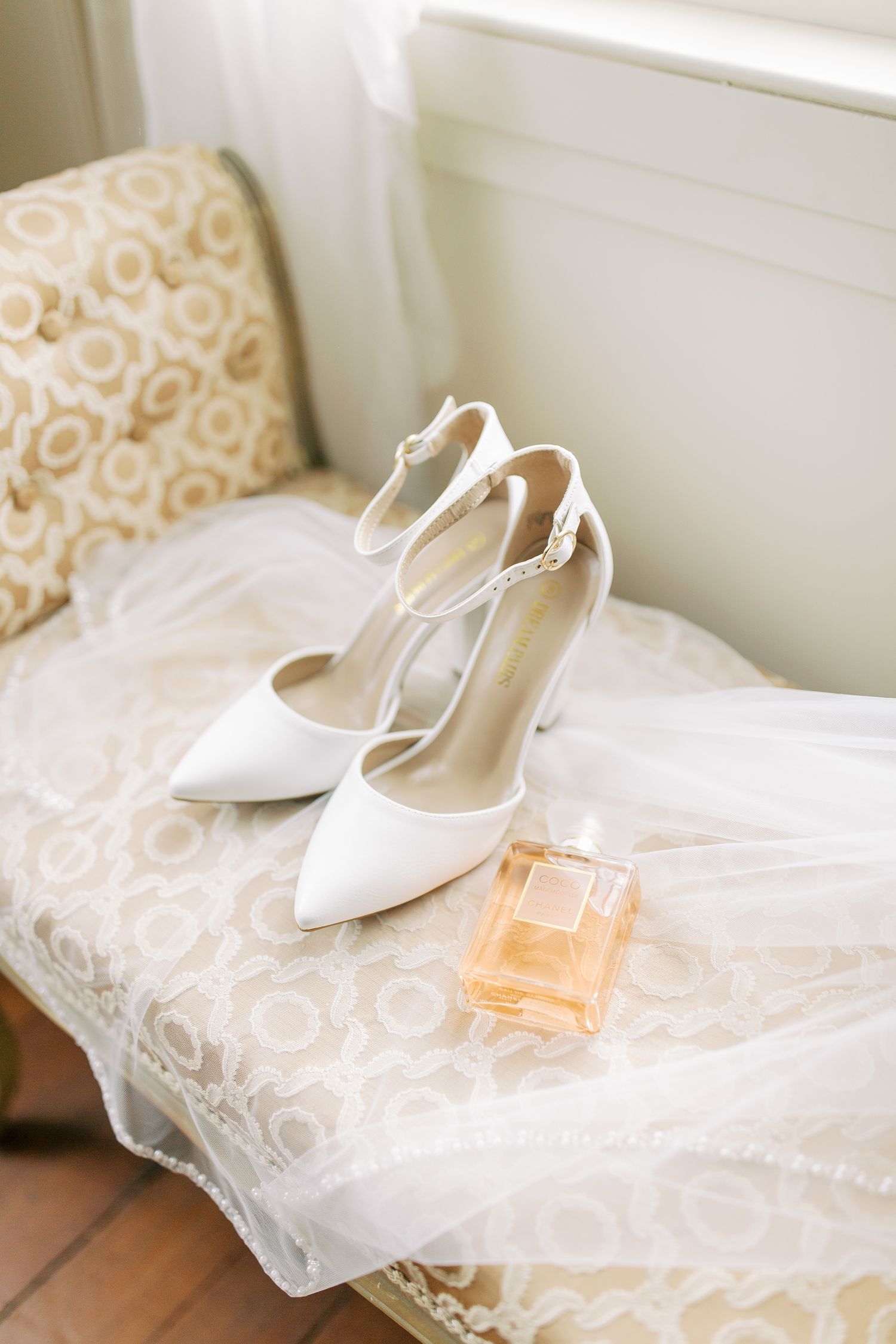 Elegant white bridal heels and perfume bottle displayed on lace-covered furniture.