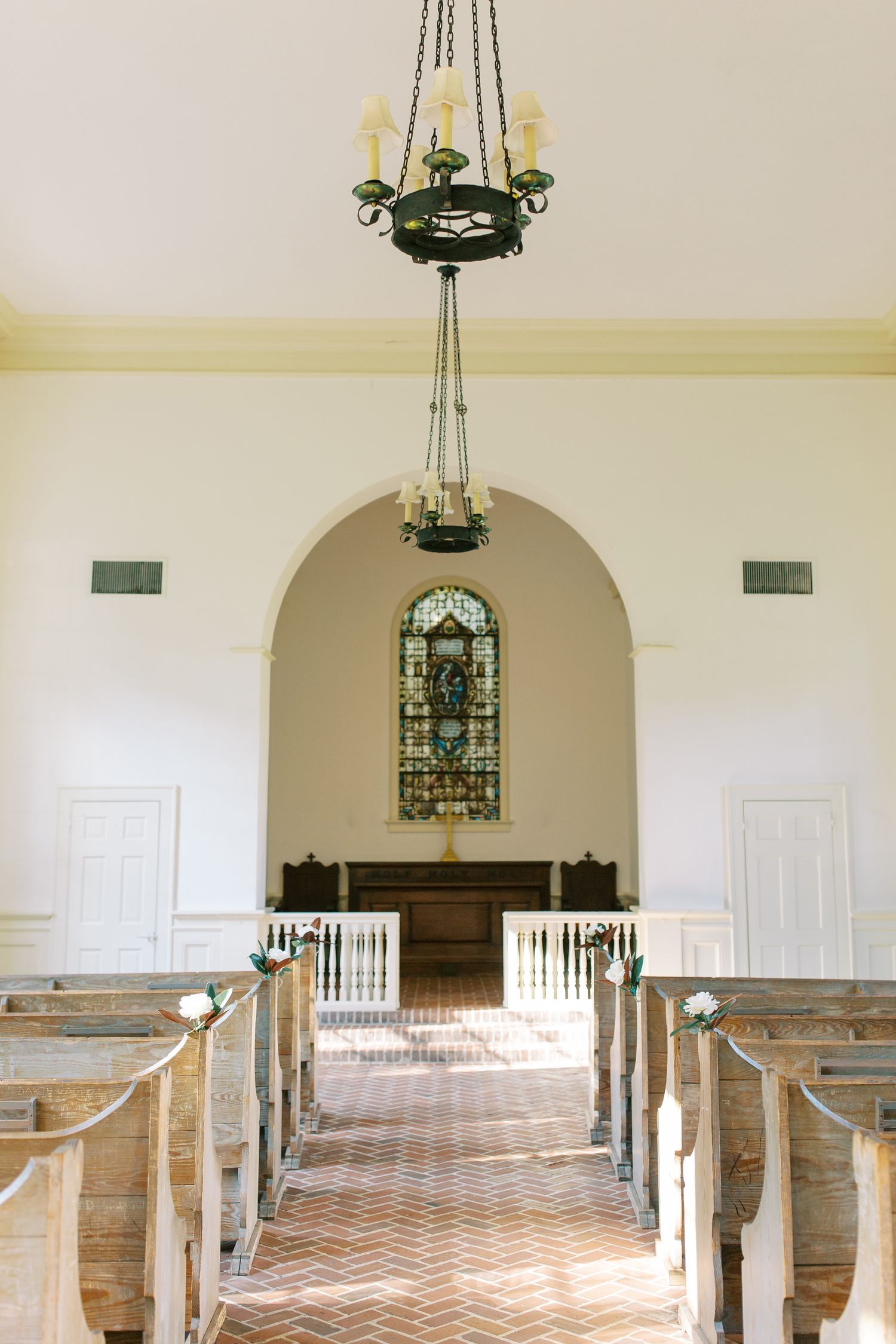 Historic chapel interior with wooden pews, chevron flooring, and stained glass window beneath chandelier.
