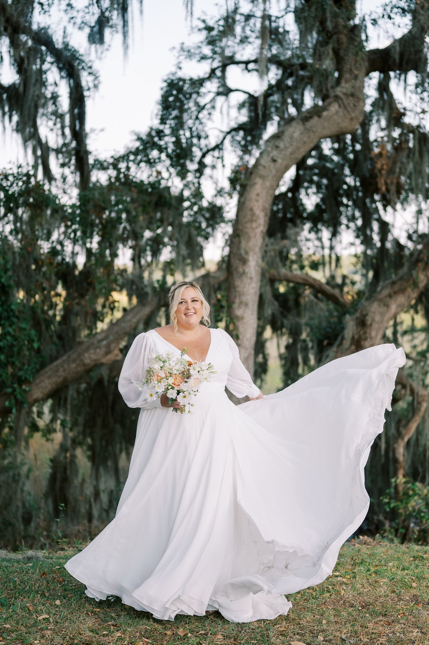 A flowing white wedding dress billows in the breeze beneath Spanish moss-draped oak trees.