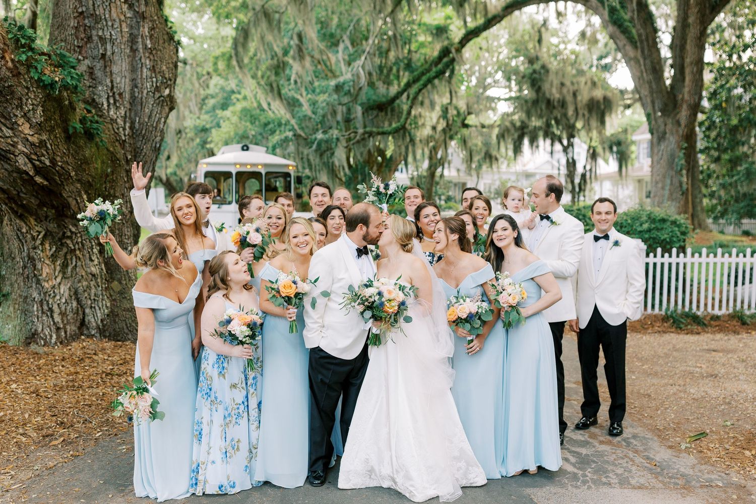 Wedding party poses beneath Spanish moss trees wearing light blue dresses and white suits.