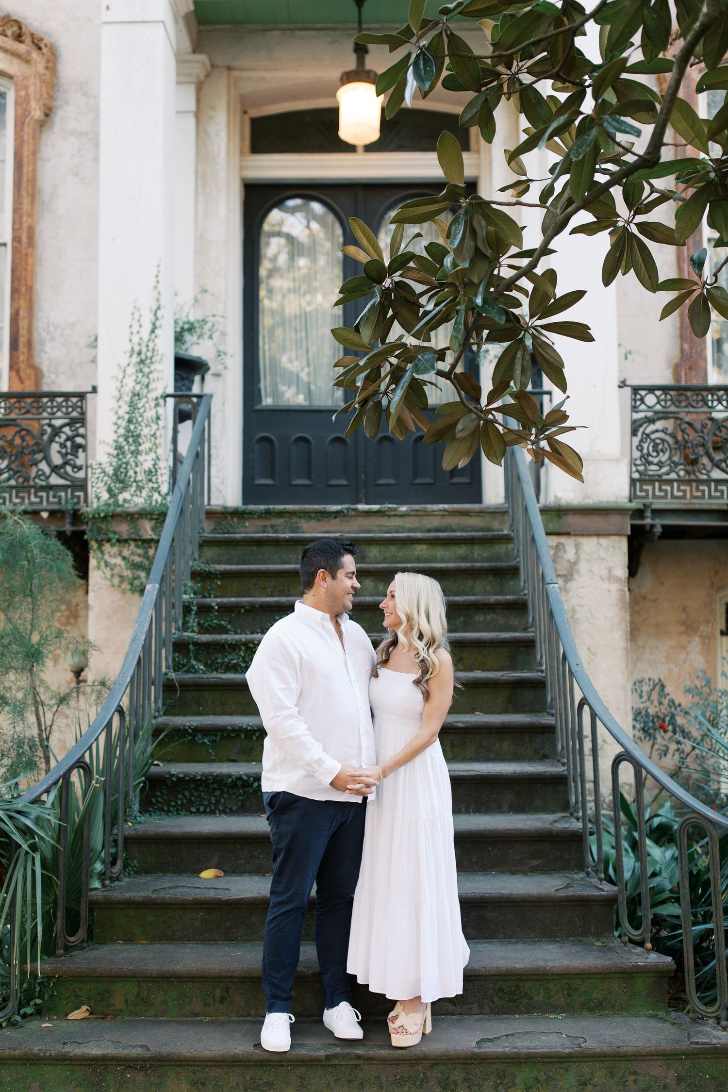 Couple in white outfits posing on vintage steps with magnolia tree in Savannah, GA