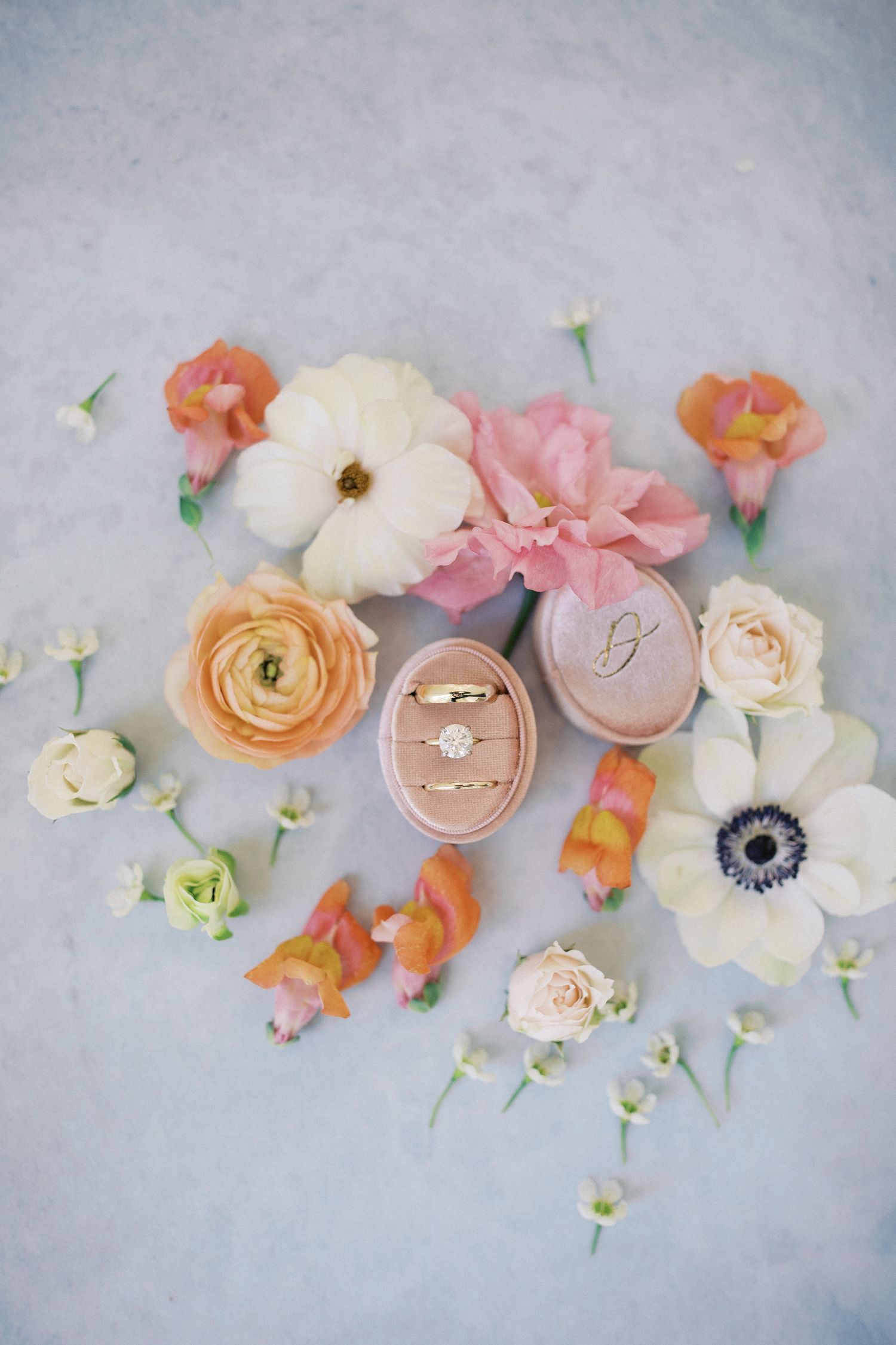 Wedding rings in a pink velvet ring box surrounded by delicate spring flowers in coral, pink and white on gray background.