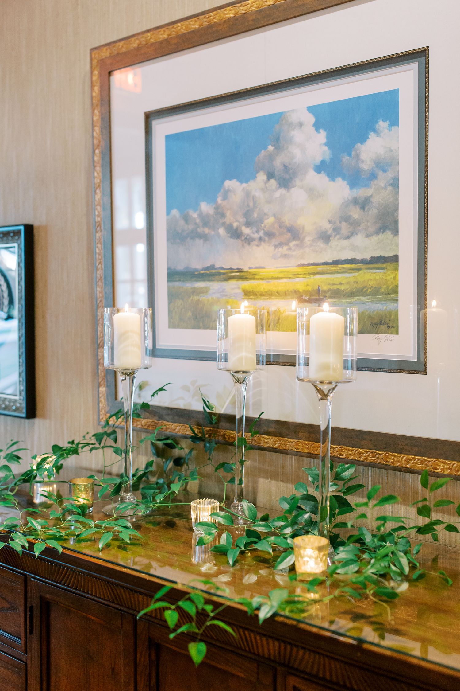 Elegant candle holders and greenery decorate a wooden sideboard beneath a framed landscape painting with clouds and marsh.