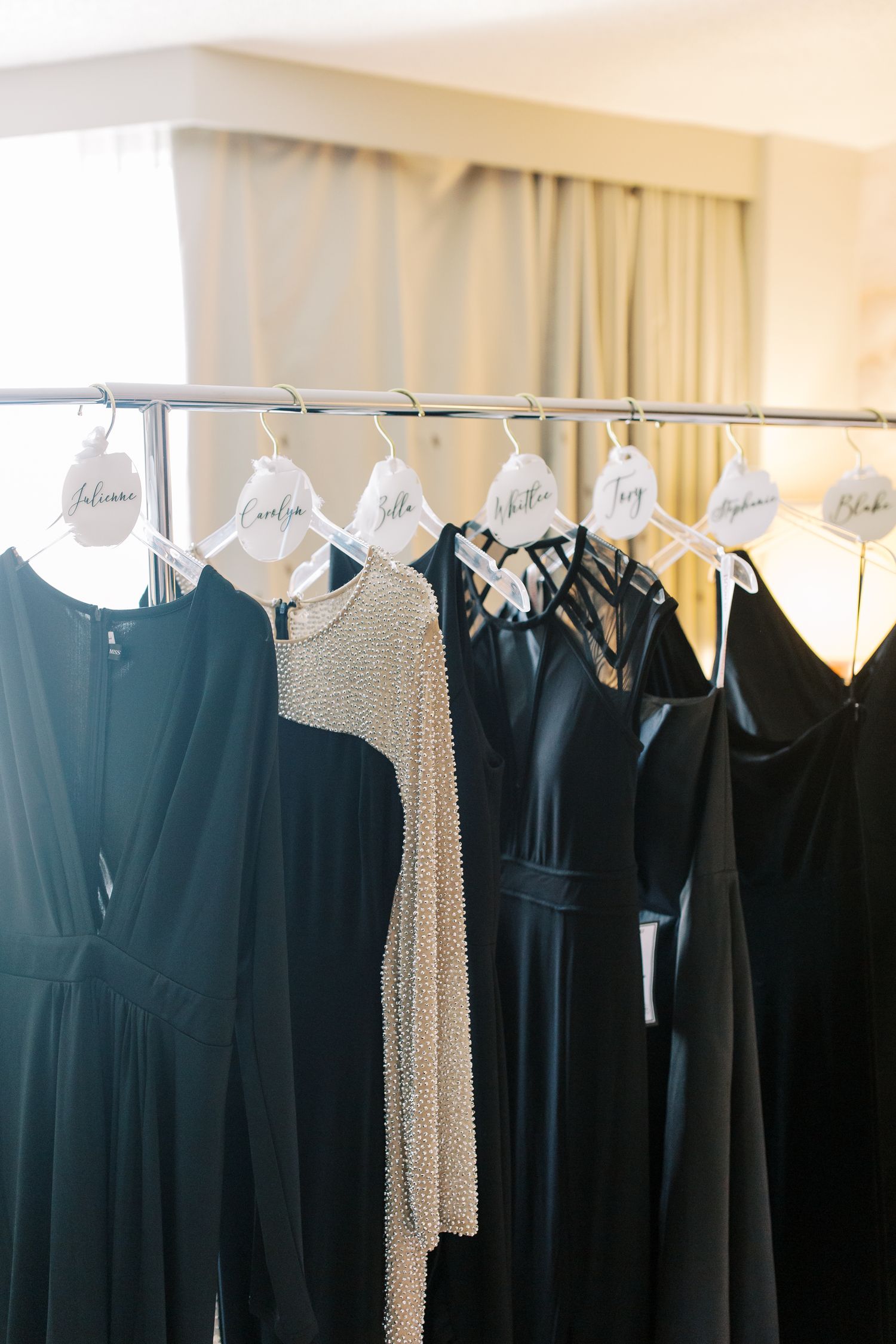 Row of bridesmaid dresses in black and sparkly beige hanging on labeled garment hangers.