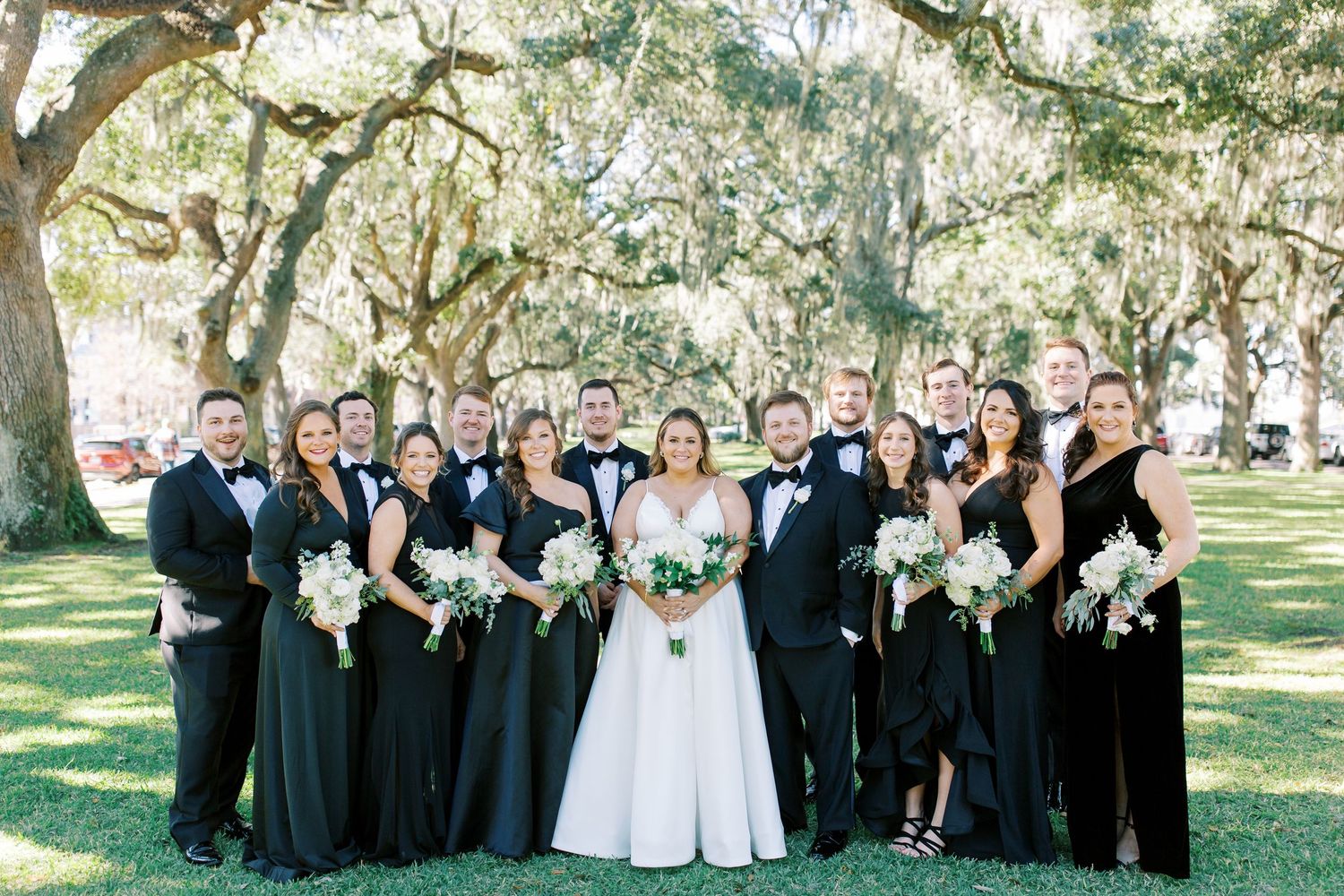Wedding party in black and white attire poses together under Spanish moss draped trees on a sunny day.