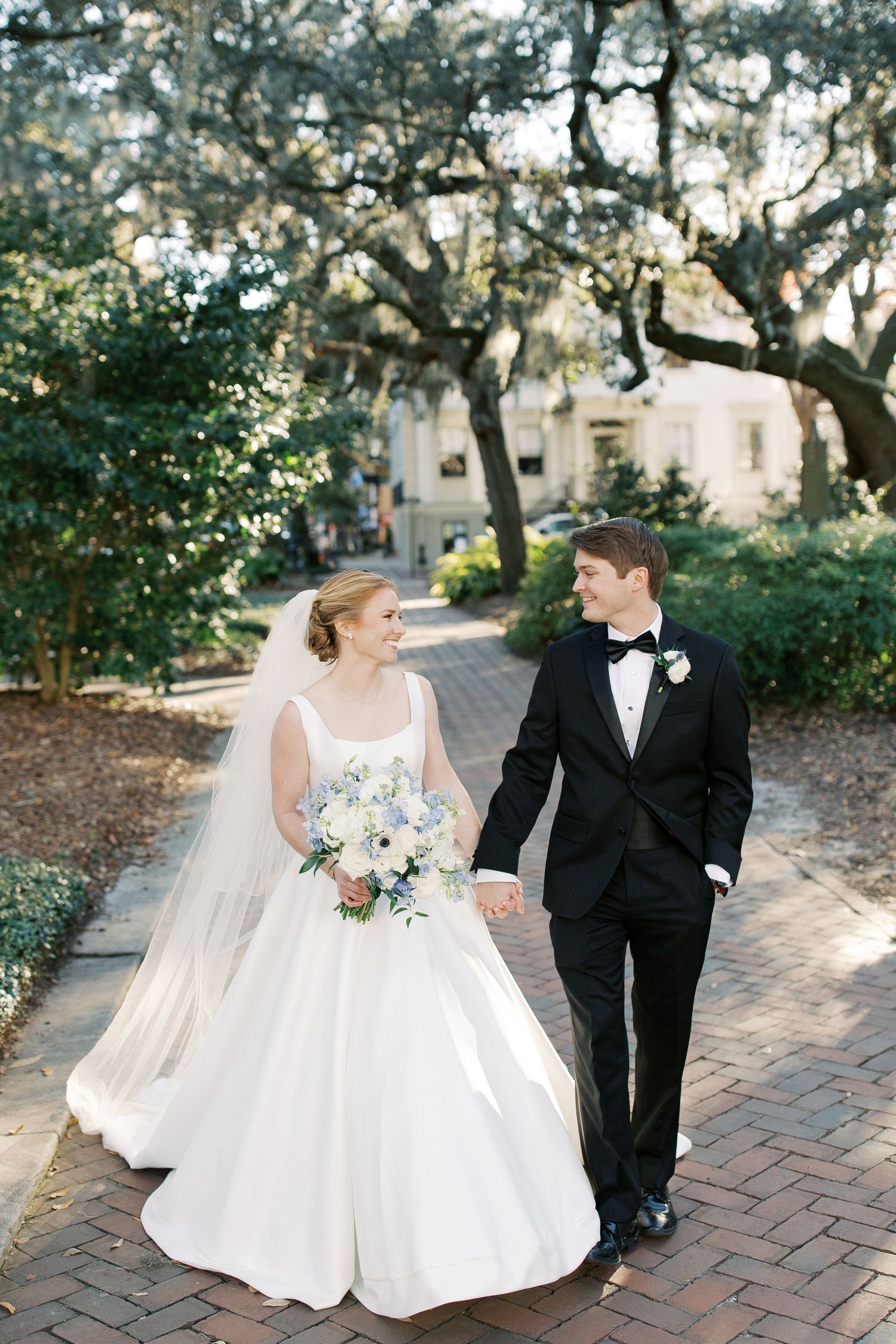 Newlyweds walking together on a brick pathway surrounded by trees.
