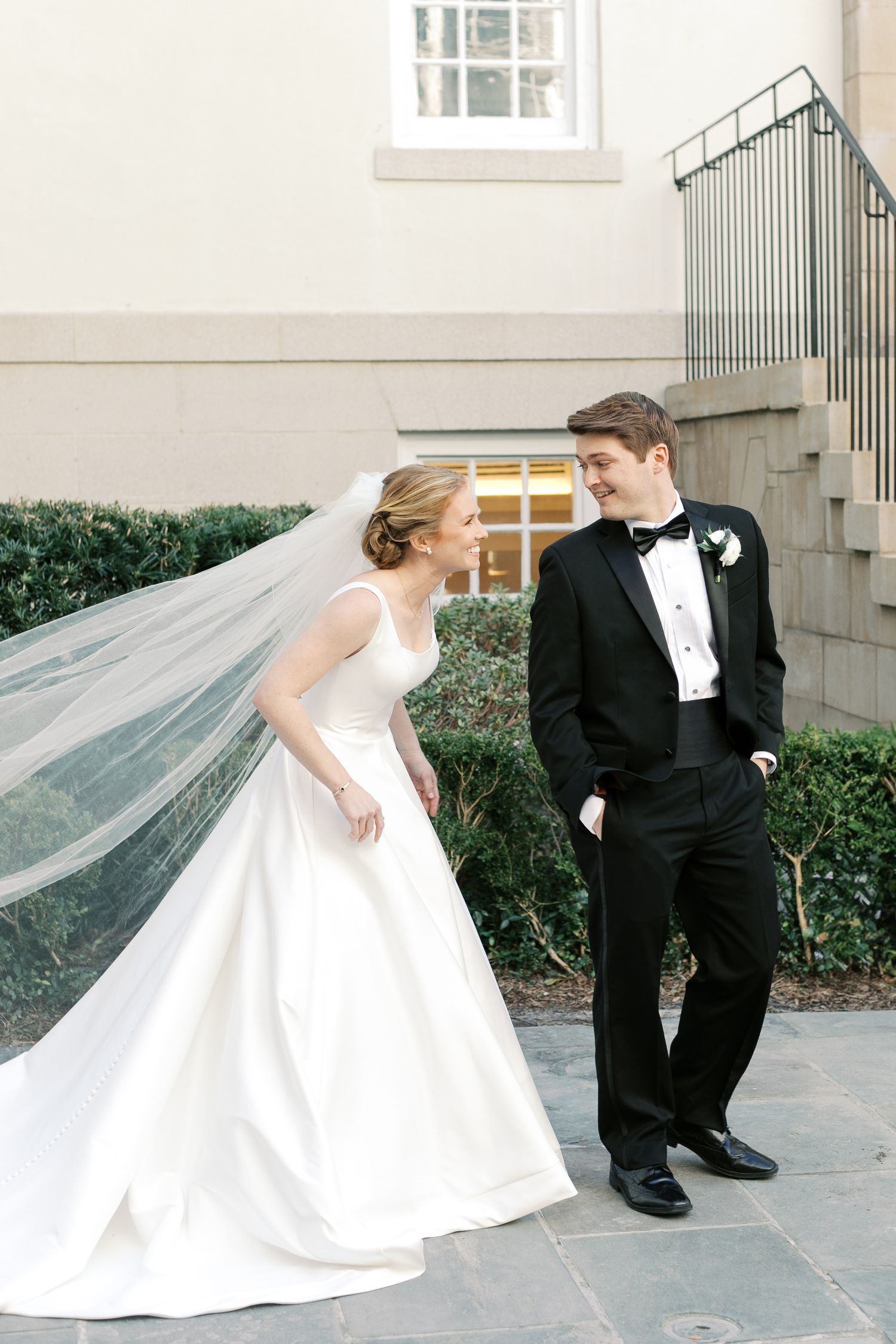 Elegant wedding couple in formal attire with flowing veil against light colored building.