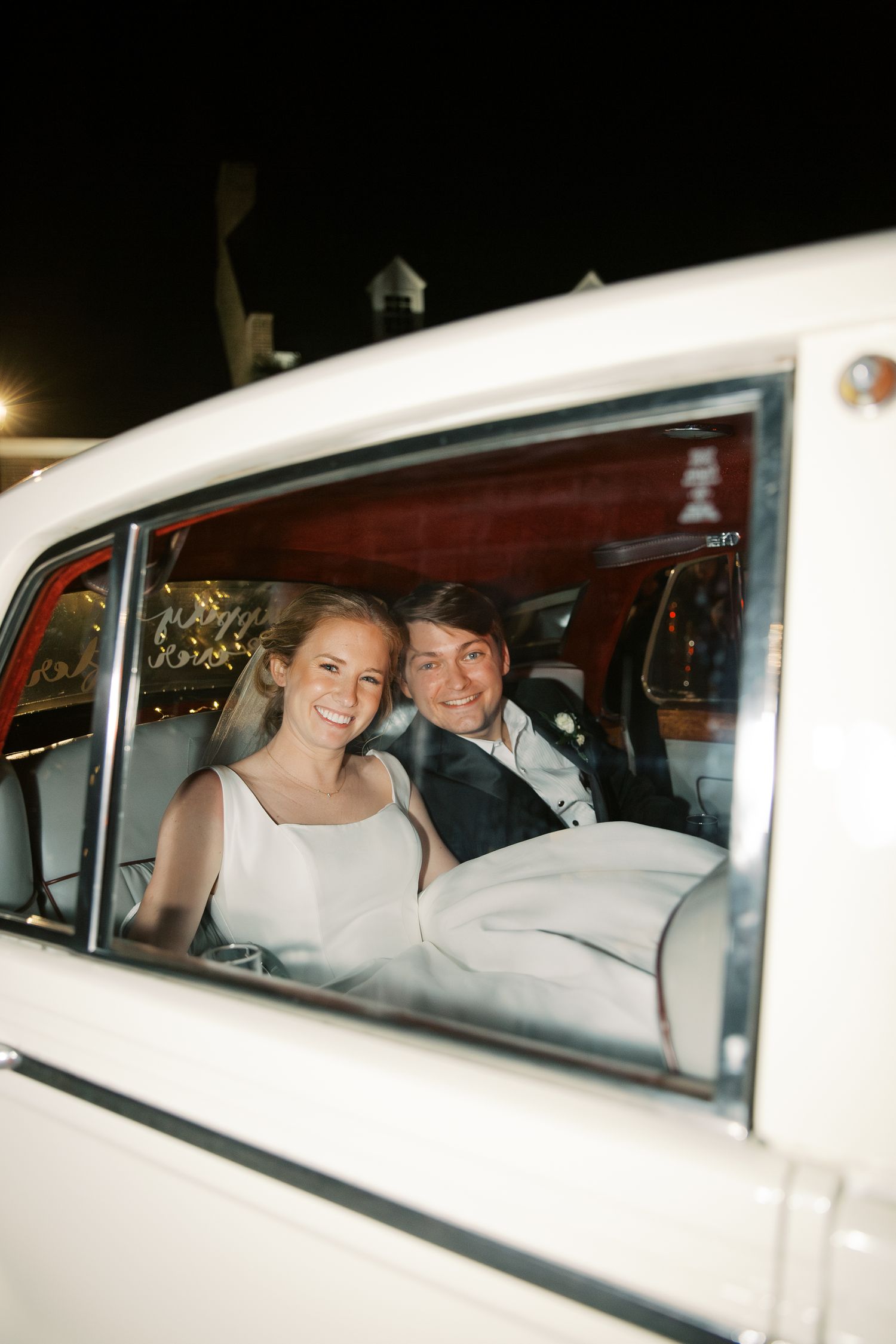 Newlyweds smiling in the back seat of a vintage white car at night.