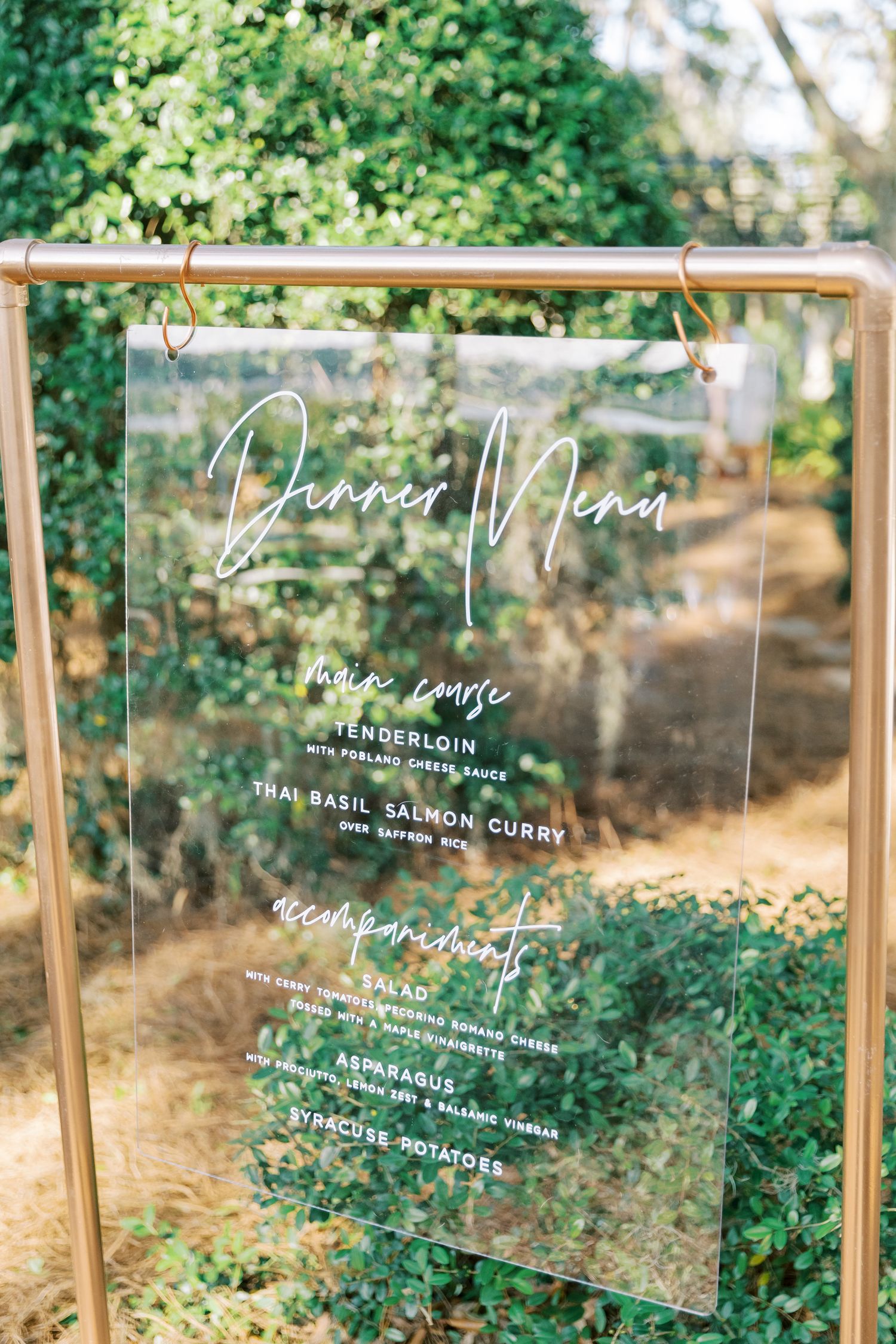 Clear acrylic wedding menu sign displayed on a gold stand with script lettering against greenery.