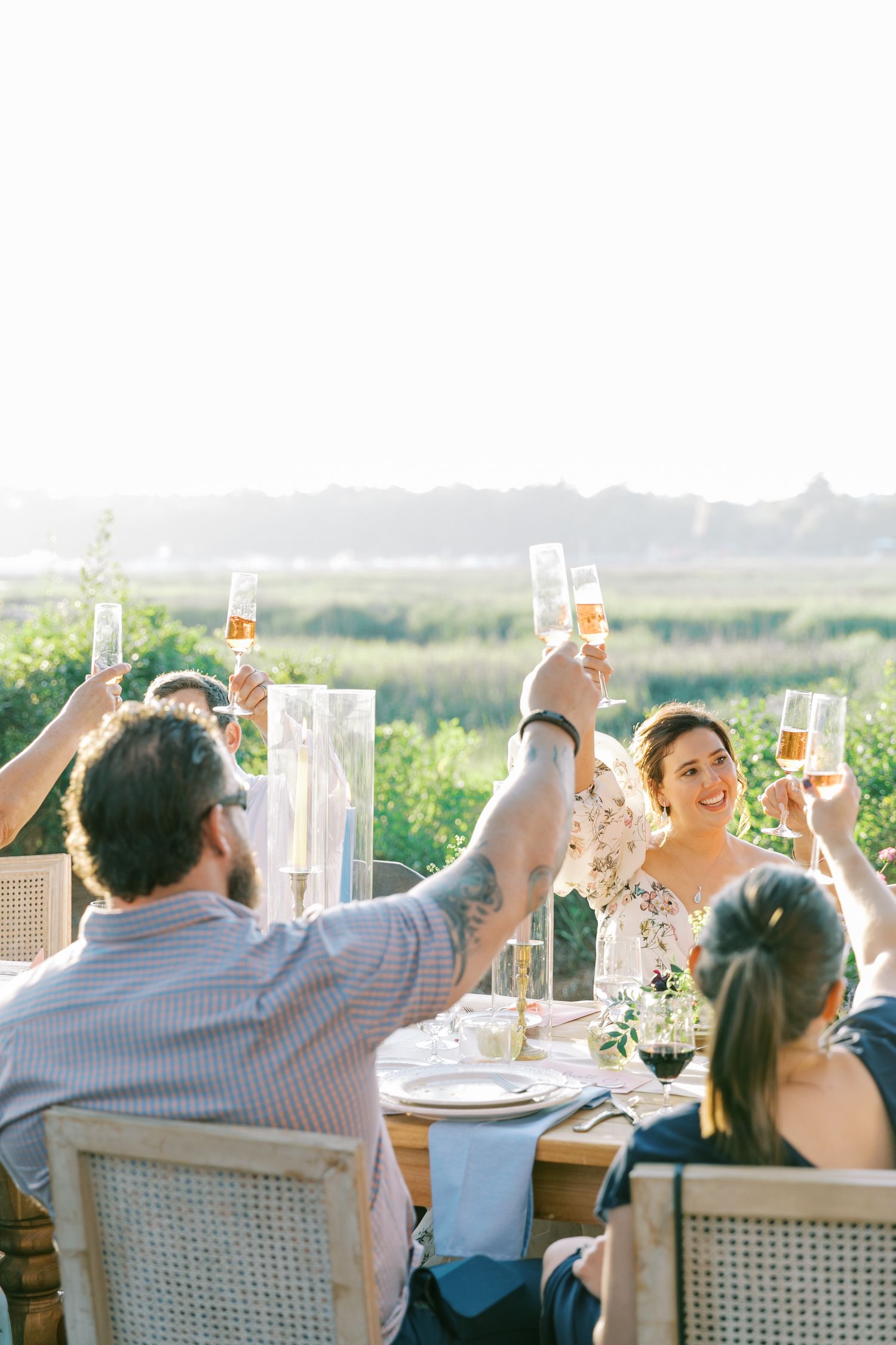 Wedding guests raise glasses in a sunset toast at an outdoor reception table.