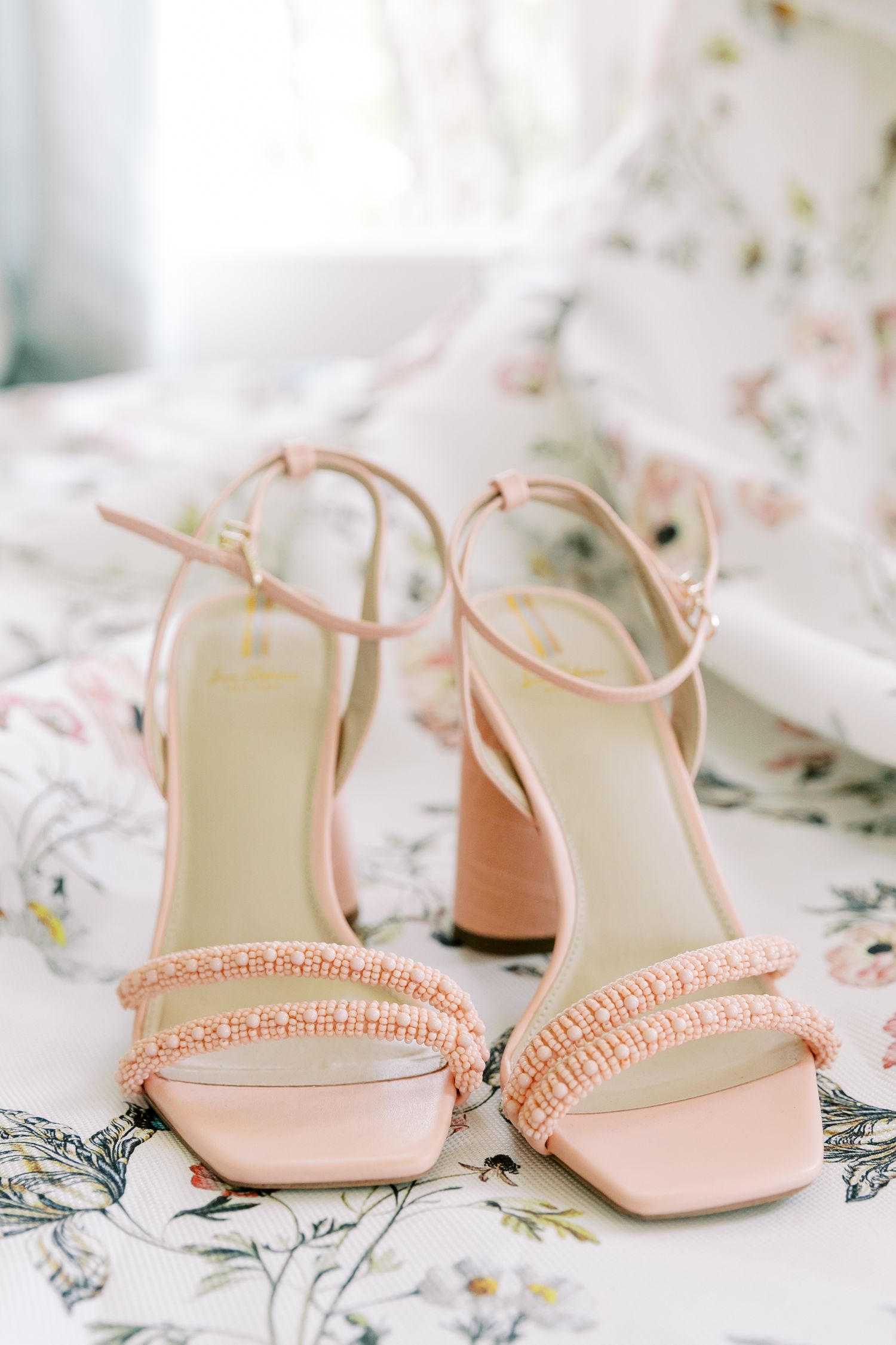 Elegant blush pink bridal sandals with crystal embellishments and block heels displayed on floral print fabric.