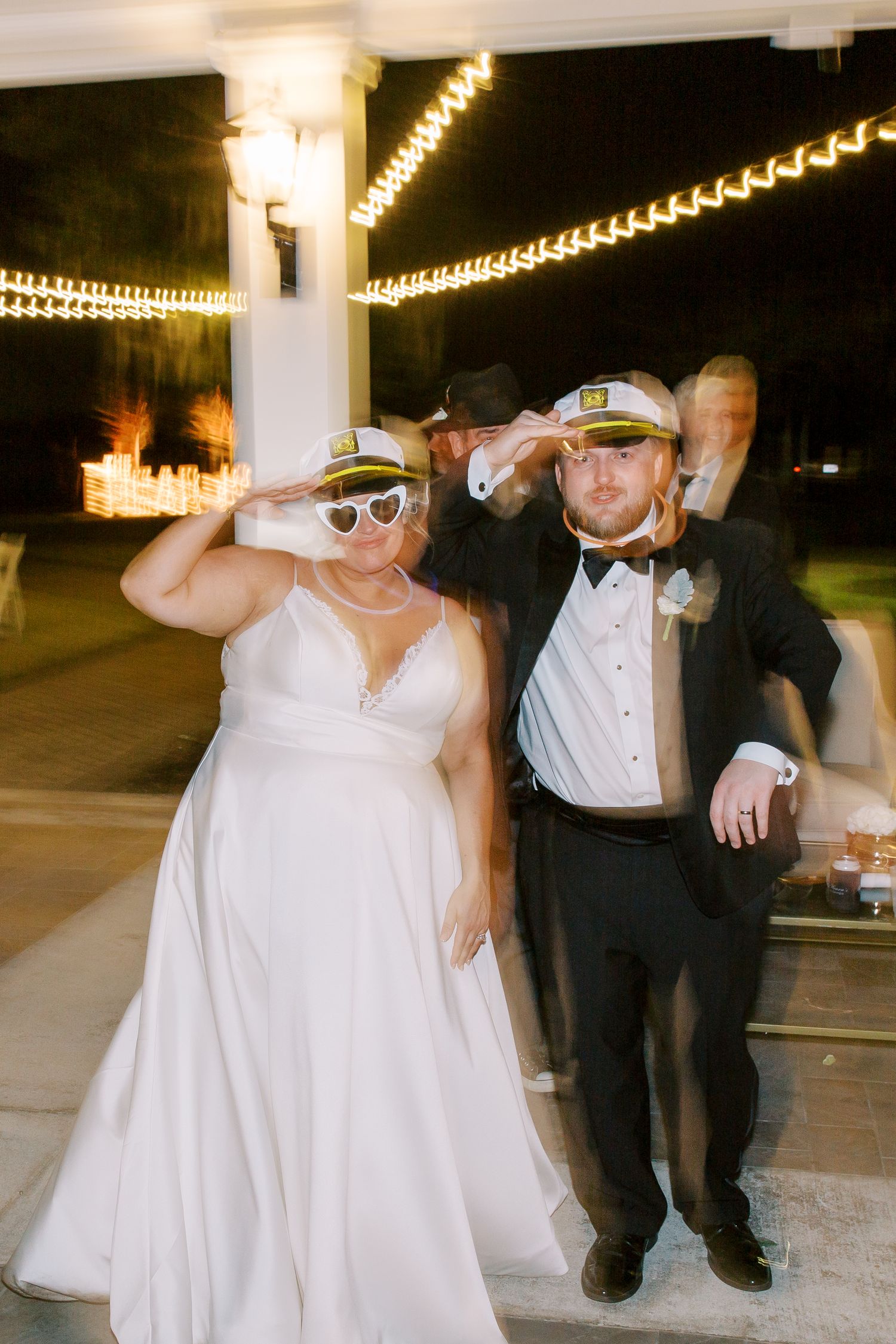 Wedding couple wearing sailor hats and sunglasses pose playfully under string lights at nighttime reception.