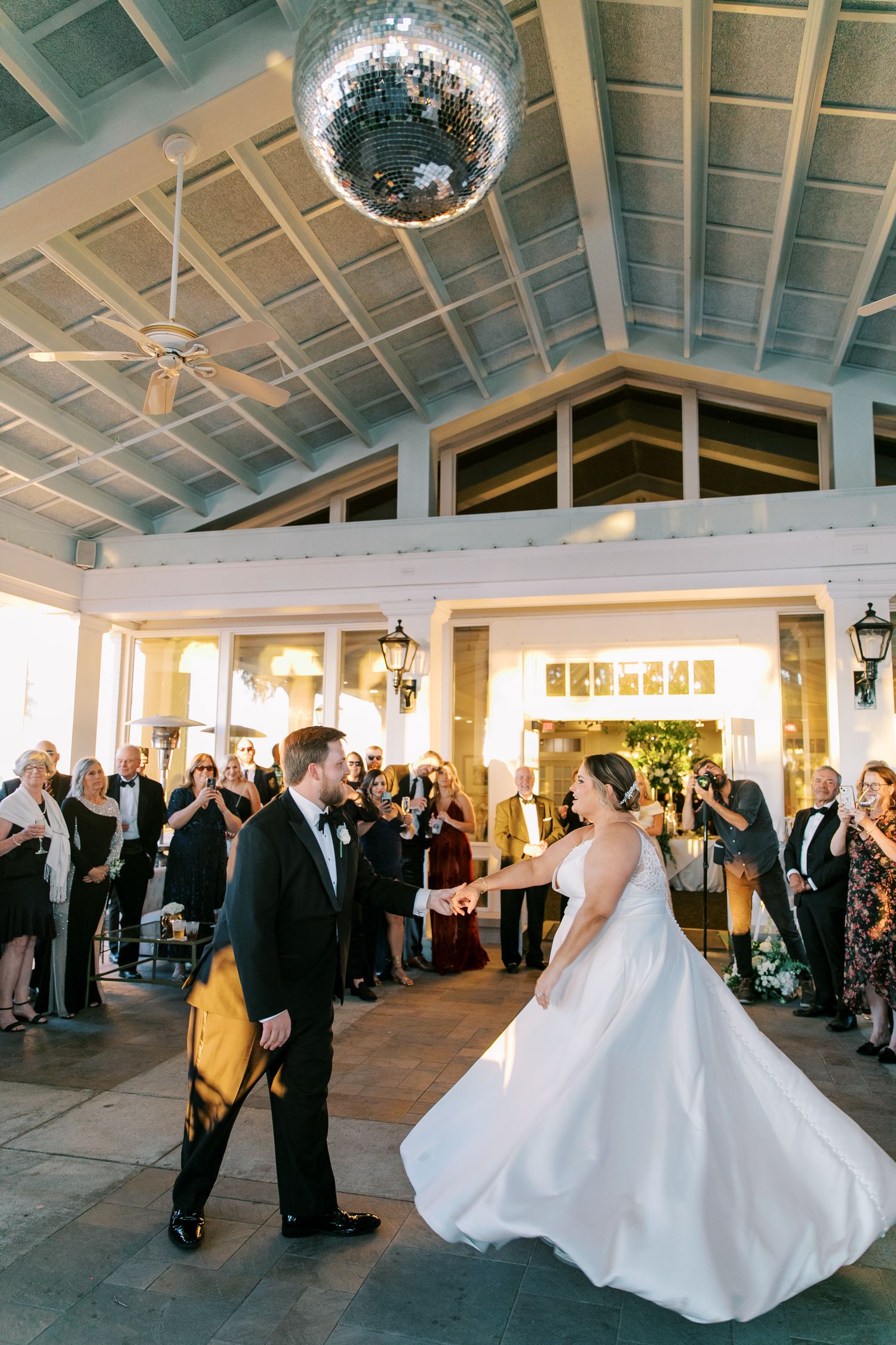 A couple shares their first dance beneath a sparkling disco ball in an elegant reception venue lit by wall sconces.