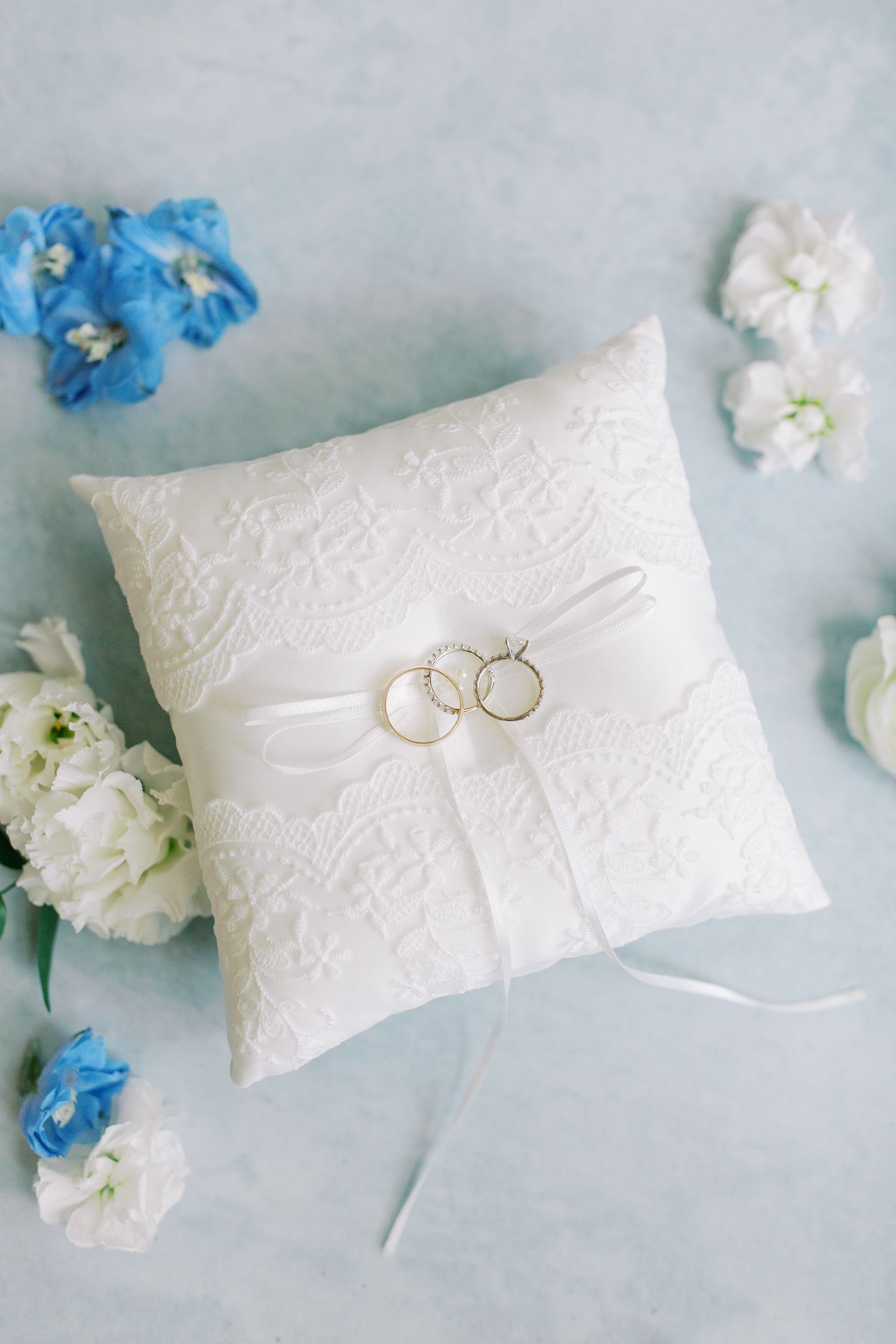 White lace ring bearer pillow with two wedding bands and delicate blue and white floral accents.