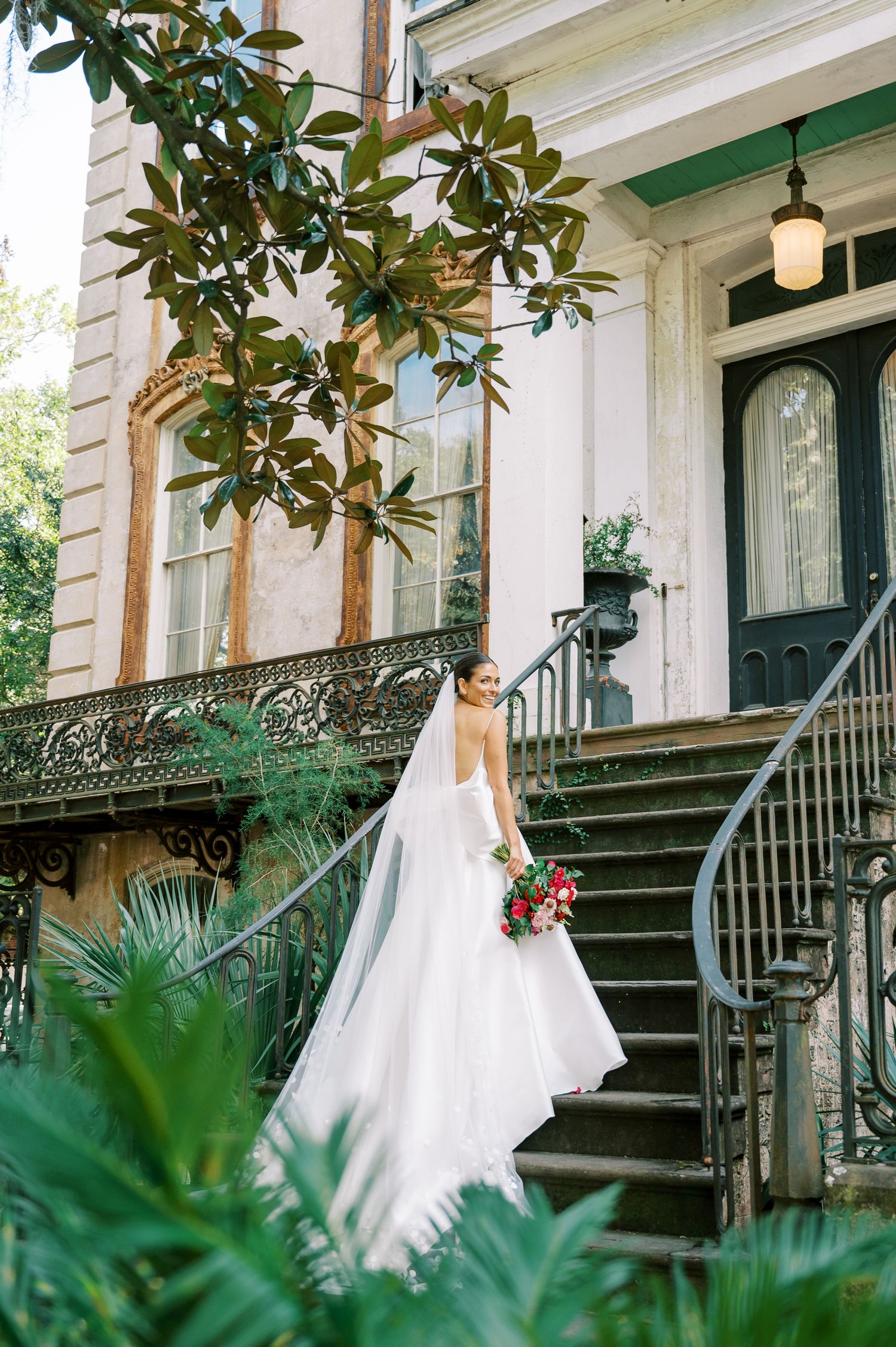 Classic bride taking portraits on historic mansion steps with magnolia and iron details.