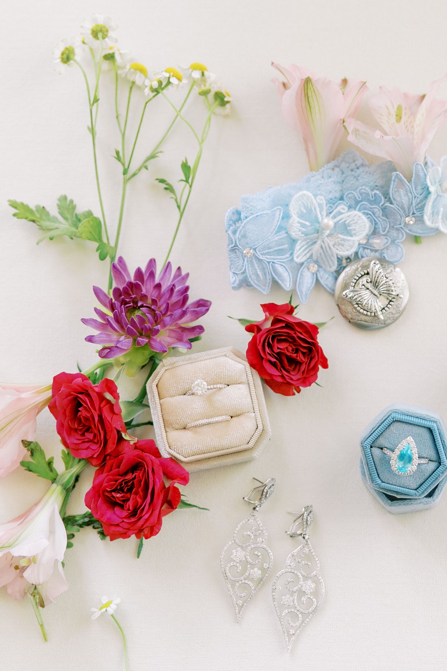 Wedding details flatlay with red roses, jewelry, and ring boxes in soft pastel colors against white background.