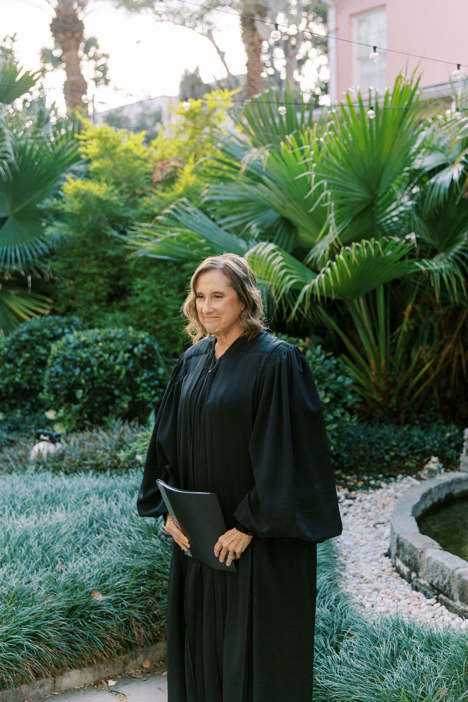 A graduate in a black academic gown stands in a lush garden setting with palm trees and tropical plants.