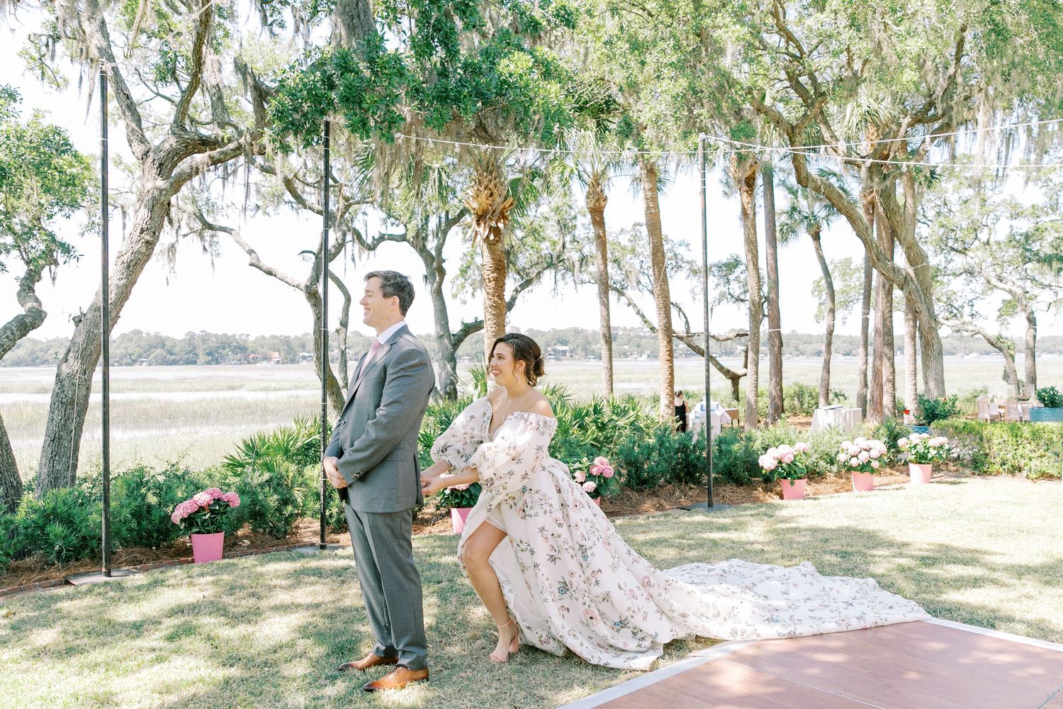 A couple in wedding attire stands in a first look moment under moss-draped trees beside a scenic marsh landscape.