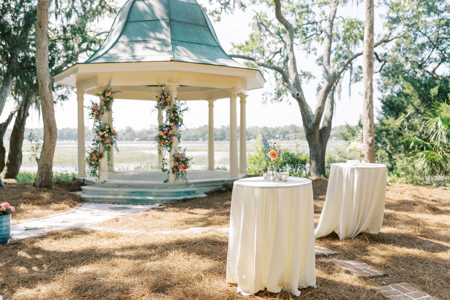 White wedding gazebos and decorative fabric drapes line a scenic tree-covered outdoor ceremony space.