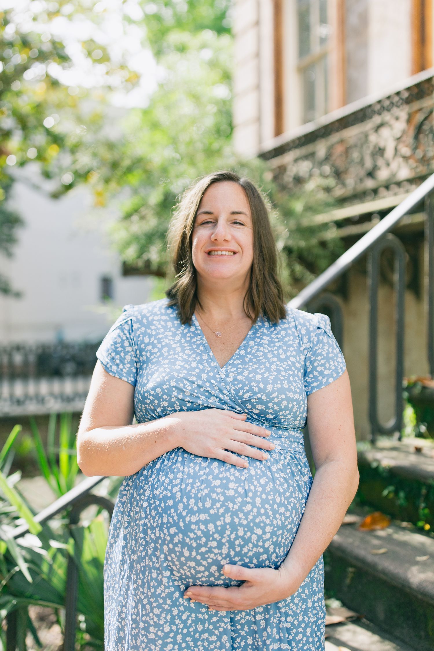 Expectant mother in blue floral dress cradles baby bump during outdoor pregnancy photoshoot.