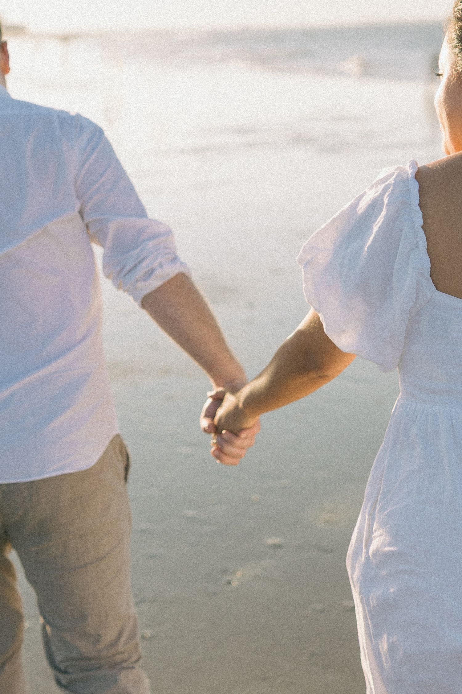 A couple in white clothing holding hands while walking along a sandy beach at sunset.