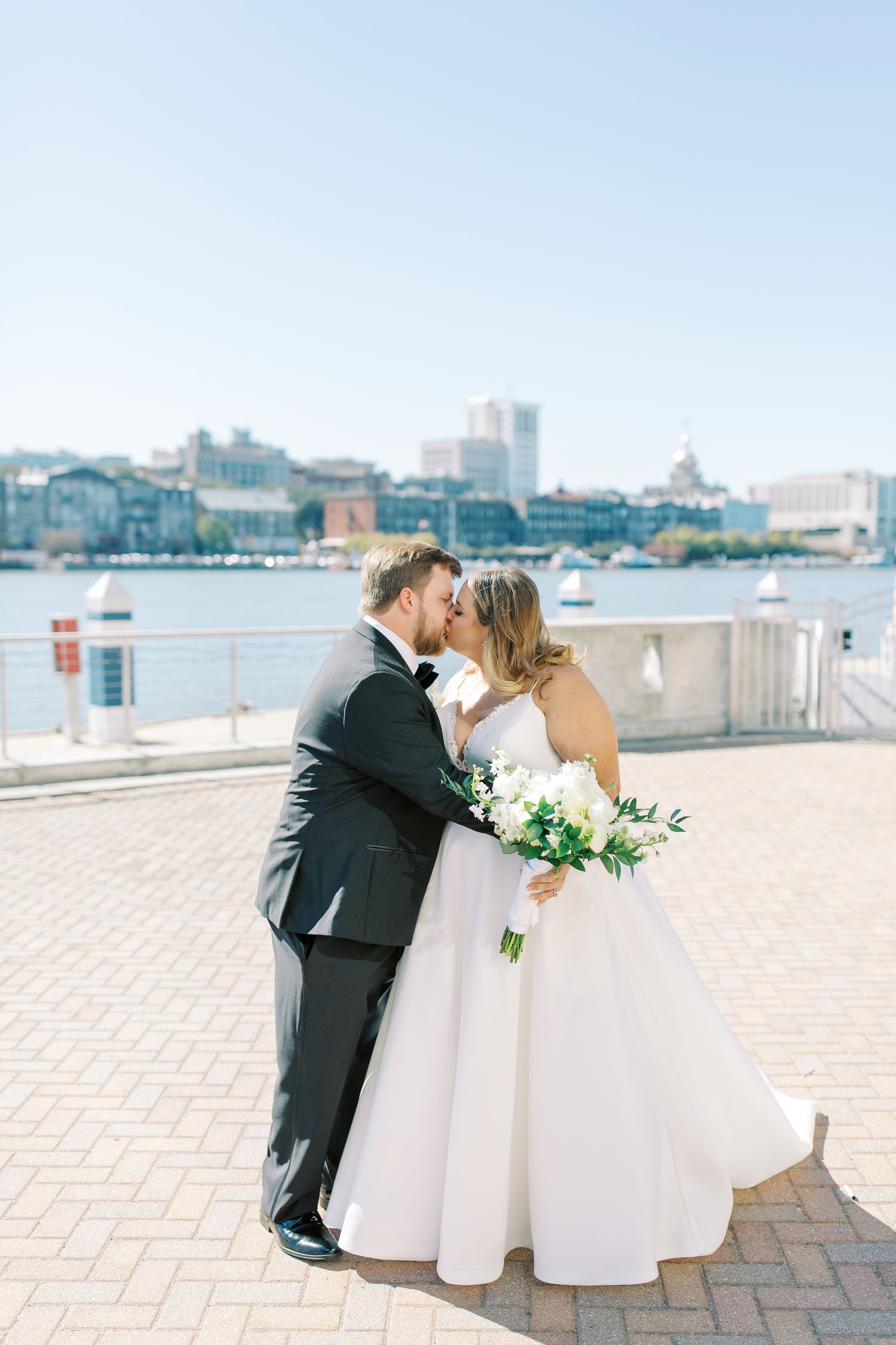 A romantic wedding couple shares a kiss on a waterfront brick patio with a city skyline in the background.