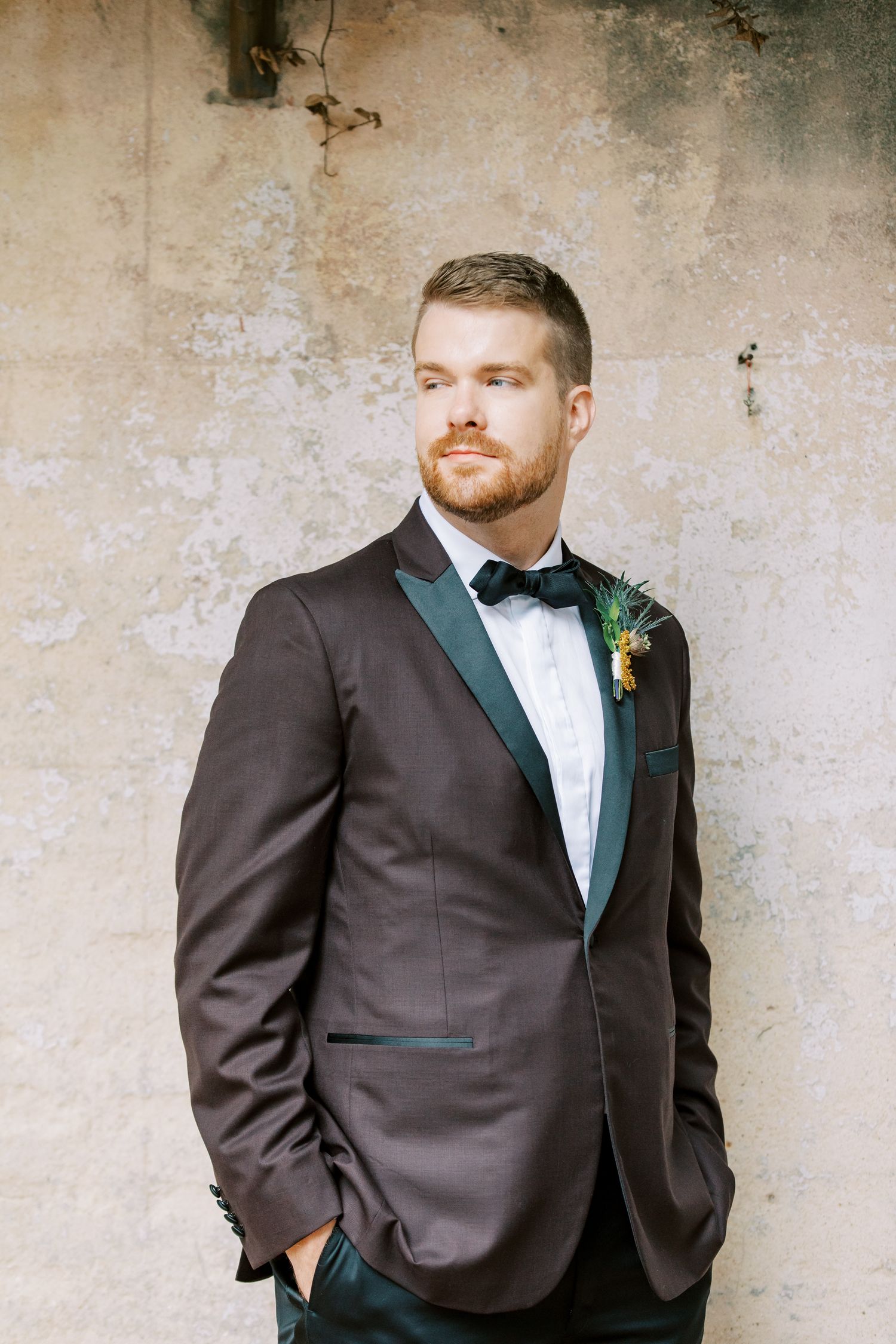 A groom in a brown tuxedo with black lapels and bowtie posing against a textured wall in downtown Savannah