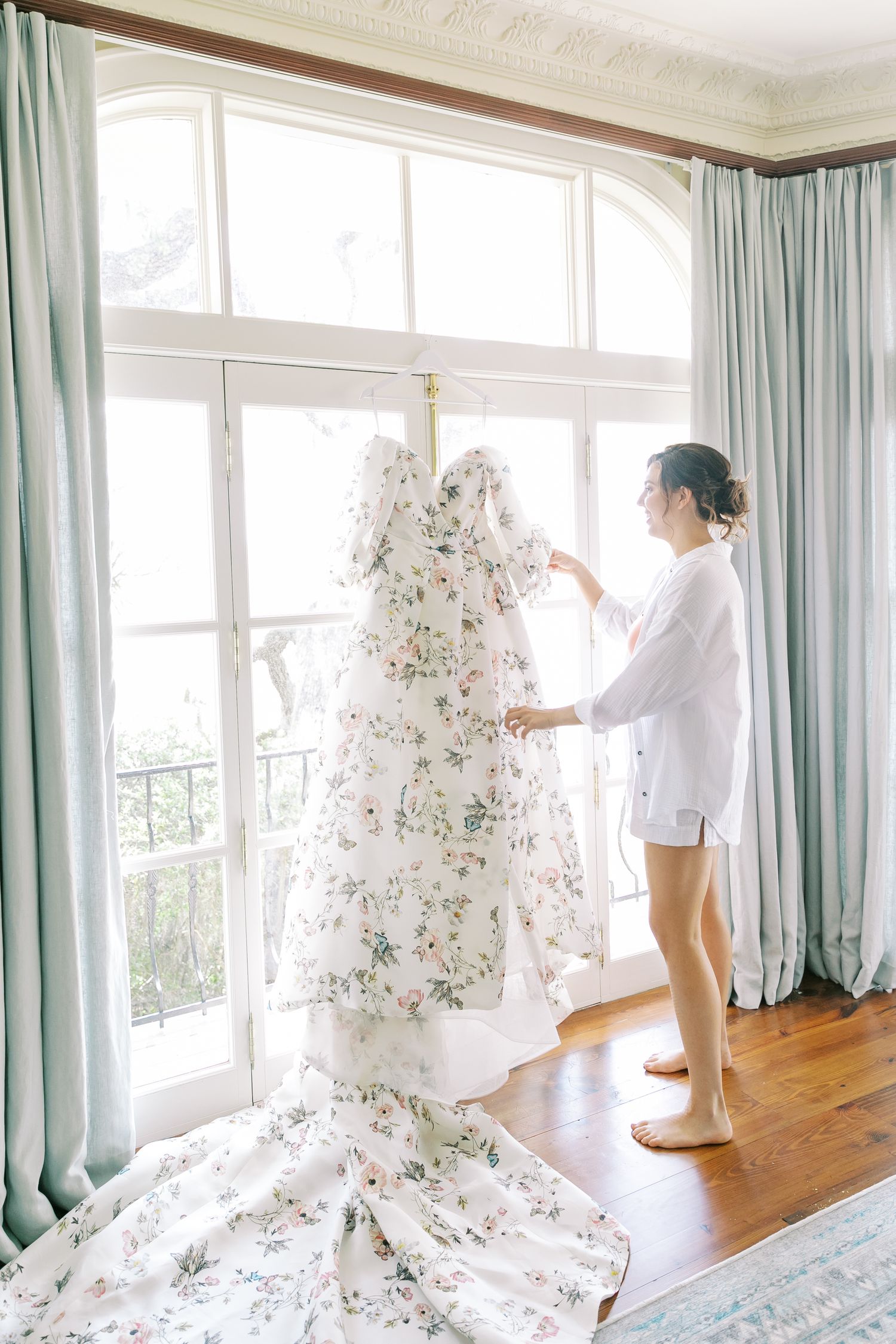 Floral wedding dress with delicate pattern hanging by large windows with seafoam green curtains.