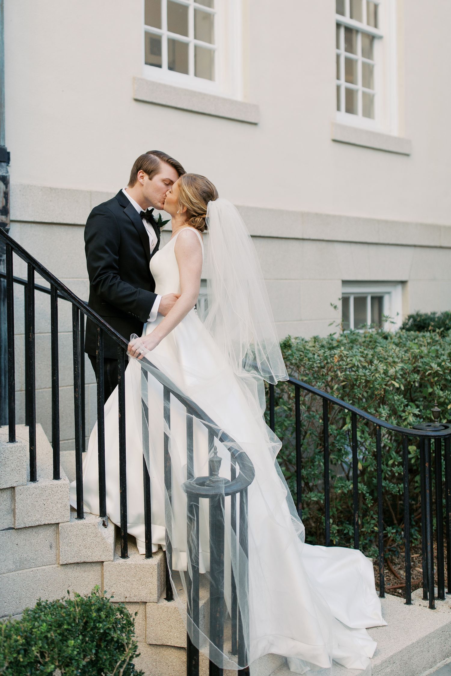 Romantic wedding couple shares an intimate kiss on stone steps with black railings against a white building.