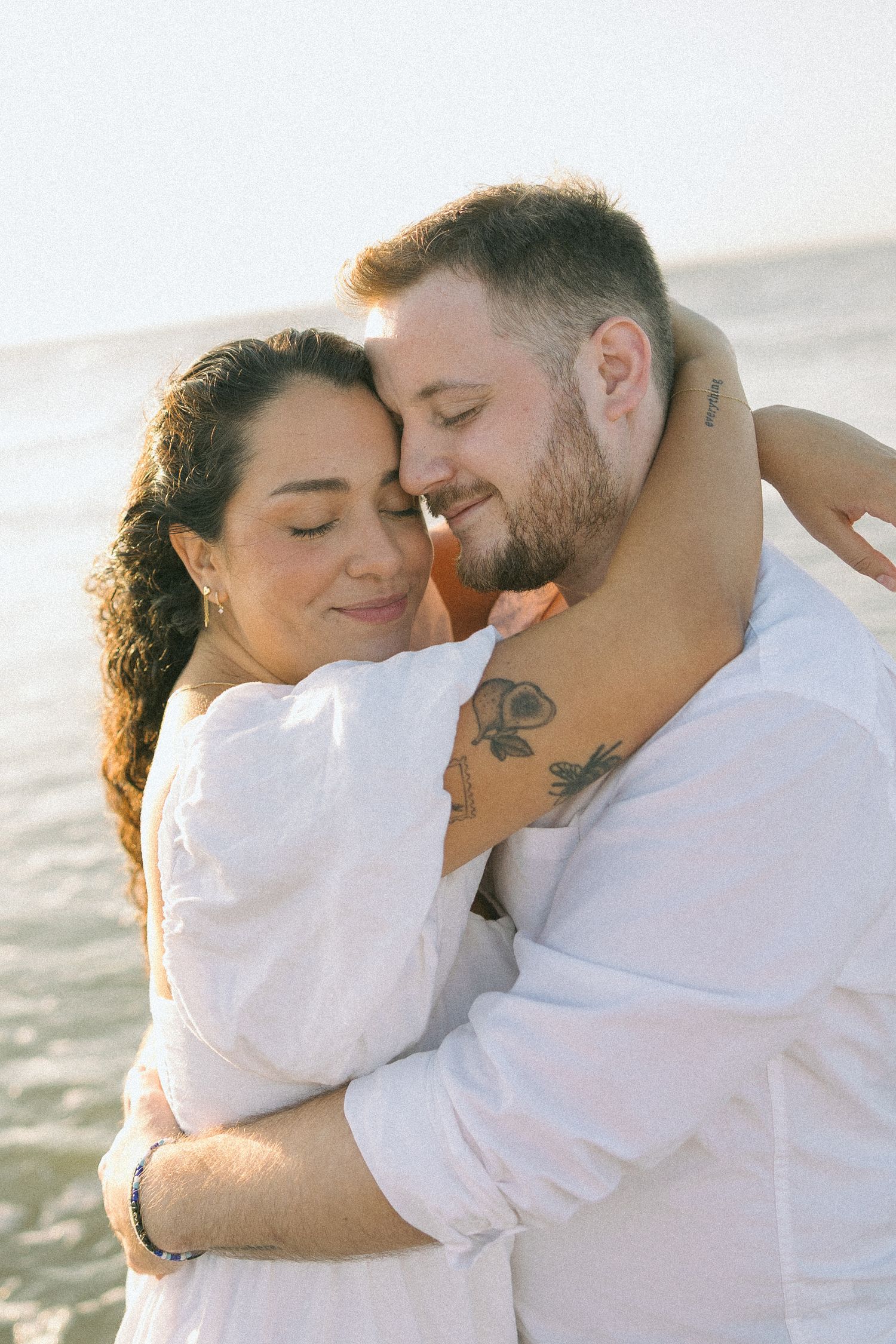 A romantic couple in white embraces tenderly on a sunny beach.