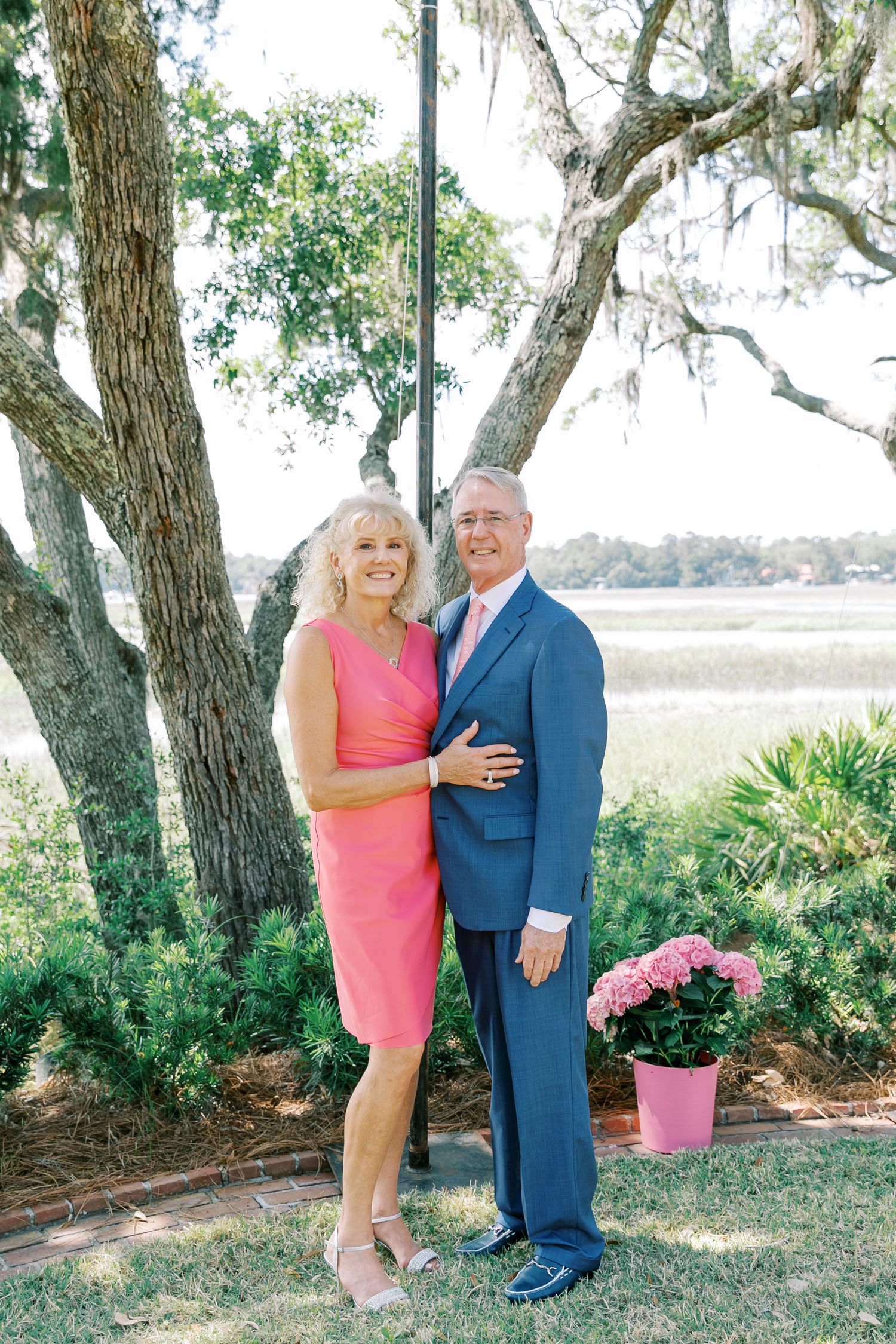 A couple in formal attire poses together under oak trees in a scenic outdoor setting.
