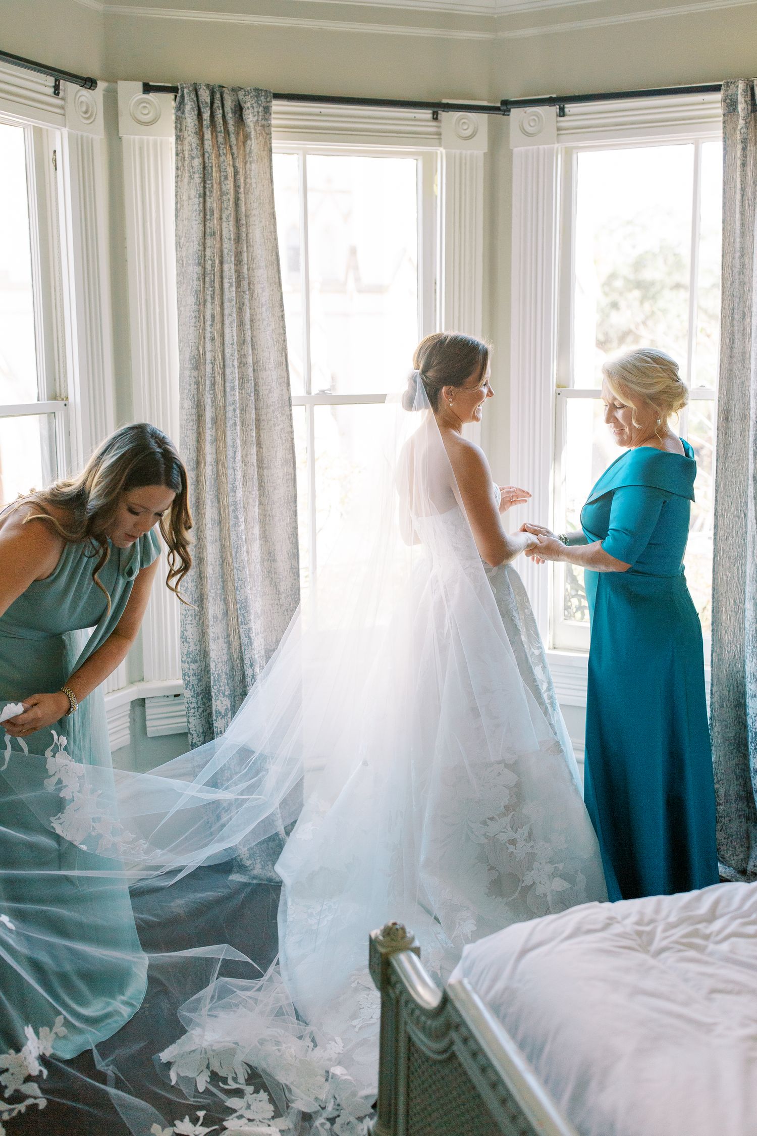 Getting ready scene with bridesmaids helping adjust a flowing wedding gown near bright windows.