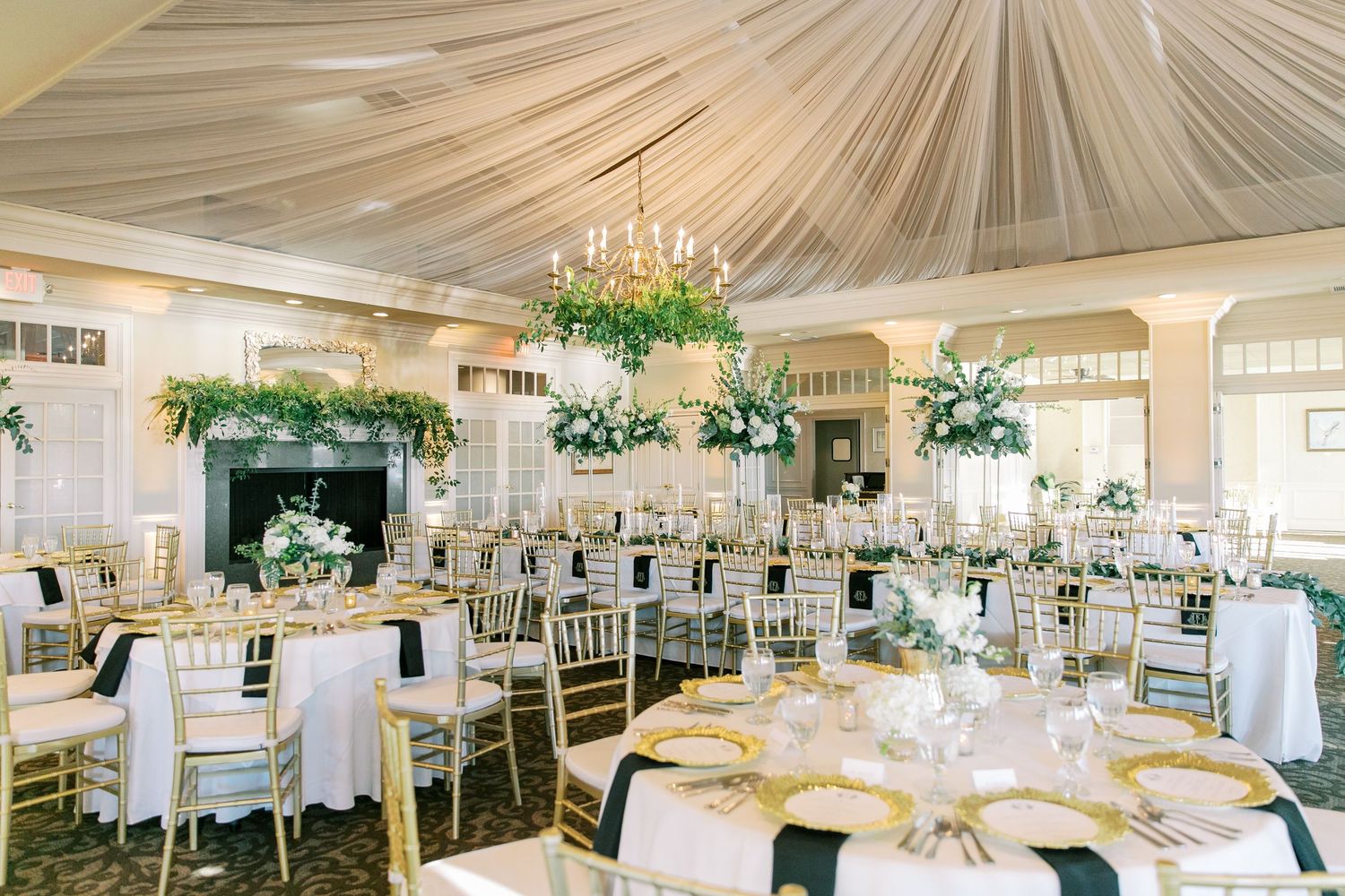 Elegant wedding reception hall with draped ceiling, crystal chandeliers, round tables, and lush green floral arrangements.