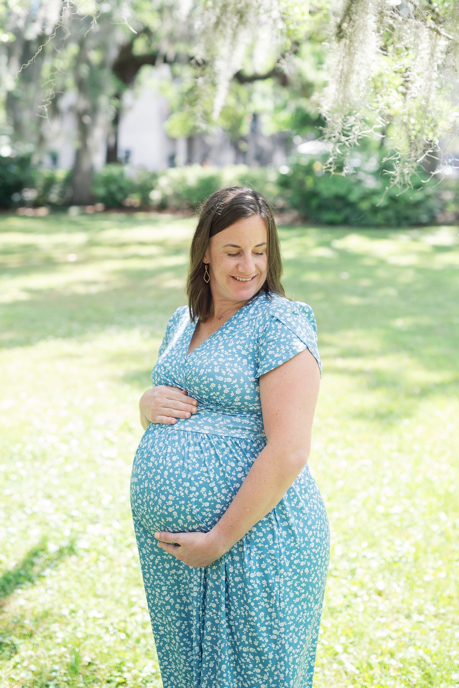 Maternity photos showing woman in blue polka dot dress posing in sunny outdoor garden setting.