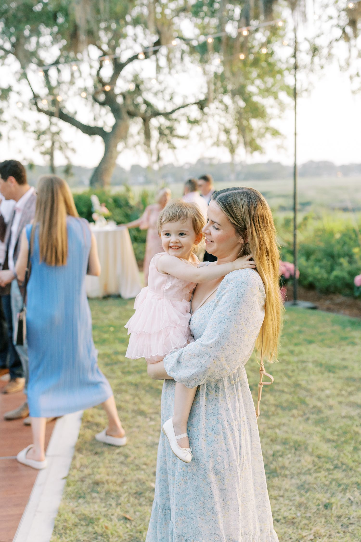 Woman in light patterned dress holds small child in pink dress at outdoor gathering as sunlight filters through trees.