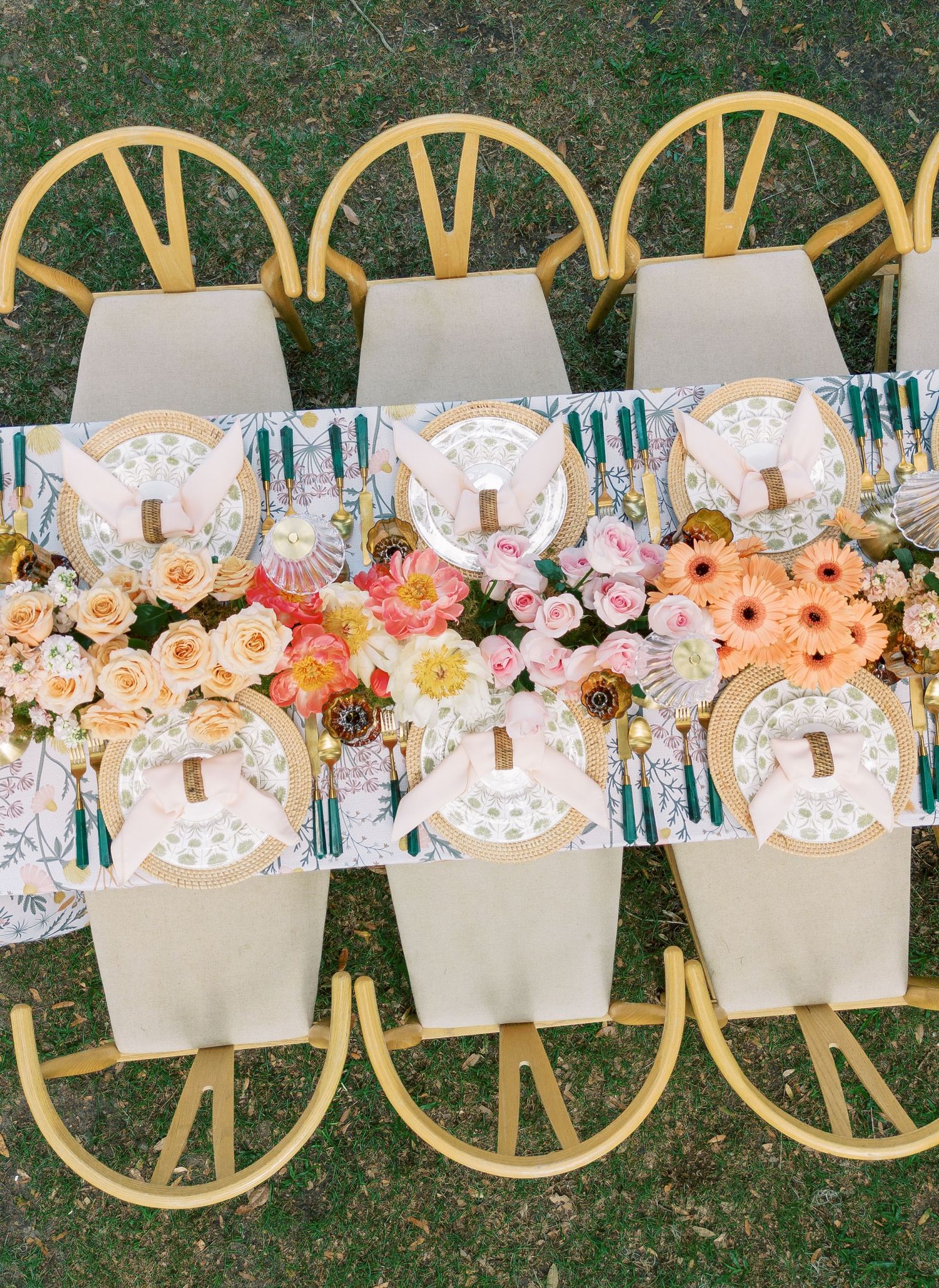 Elegant table setting with white linens and pink floral centerpieces arranged in a long row on grass for outdoor dining.