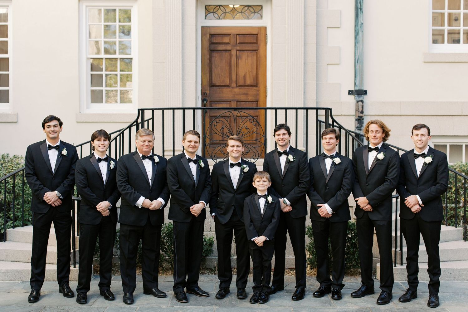 Groomsmen in black tuxedos lined up against white building exterior.