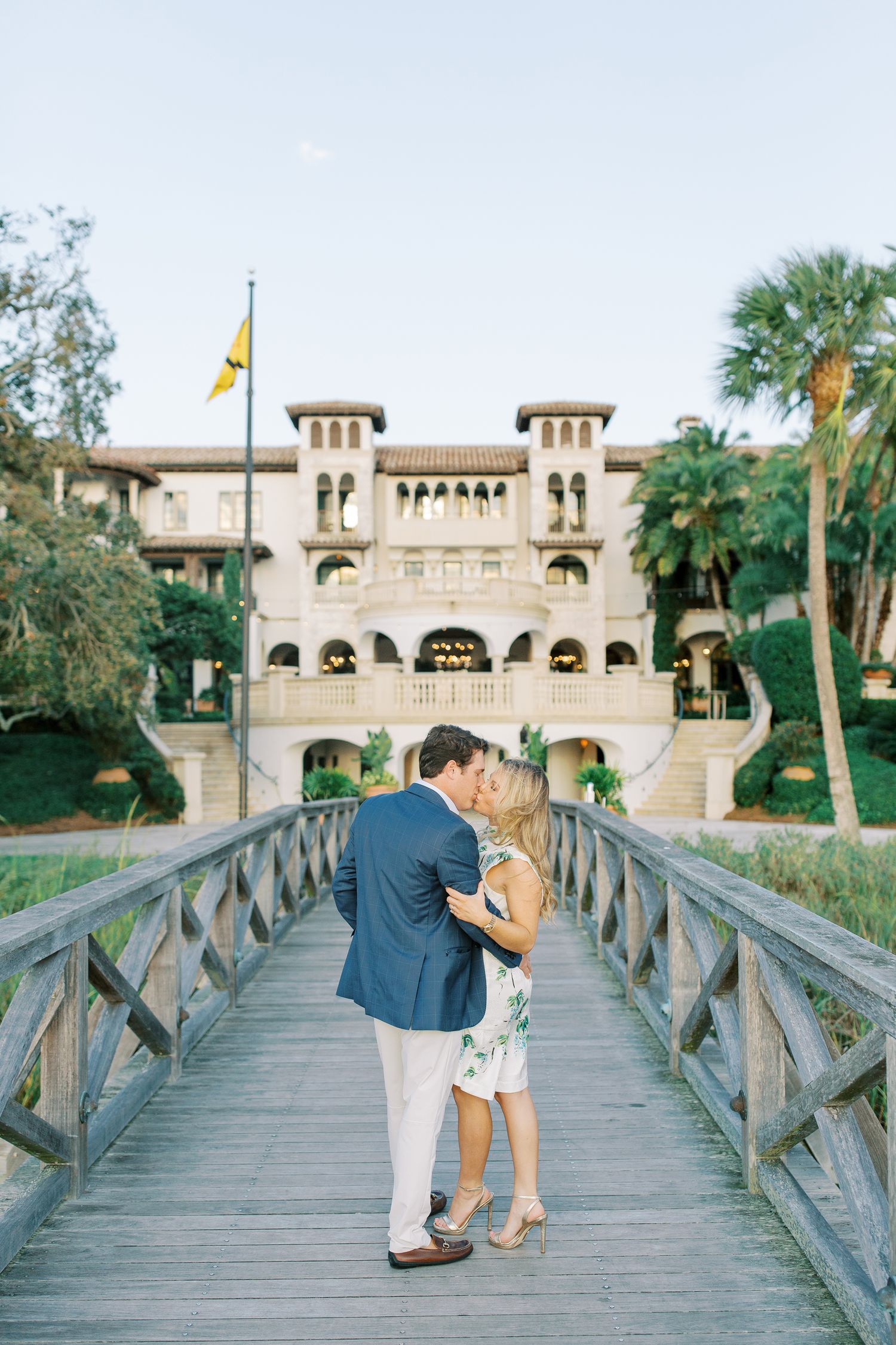 A romantic couple shares a kiss on a wooden bridge leading to a Mediterranean-style mansion with palm trees in Sea Island, GA