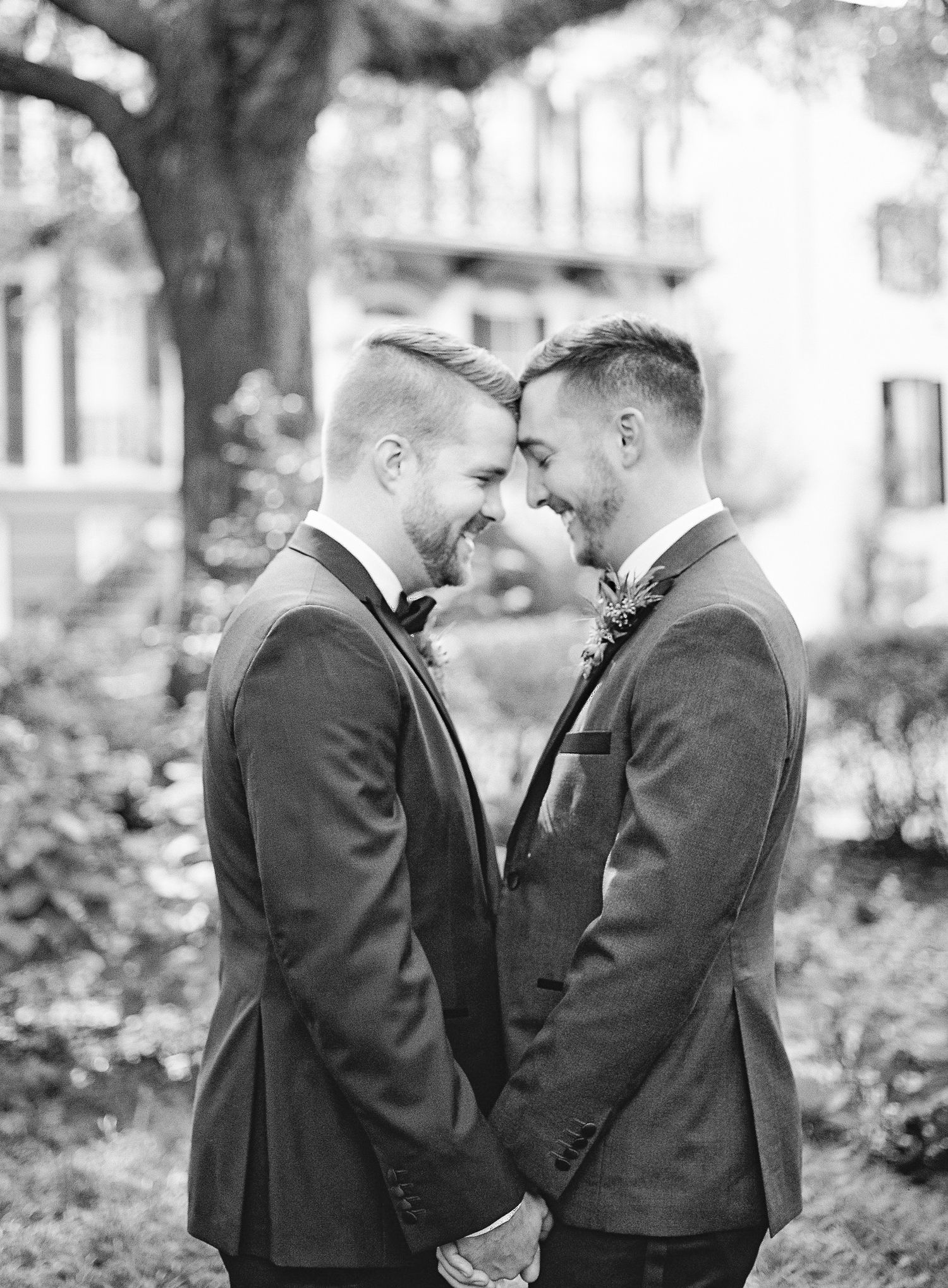 Two grooms in matching black tuxedos share an intimate moment outdoors in black and white photography in Savannah, GA
