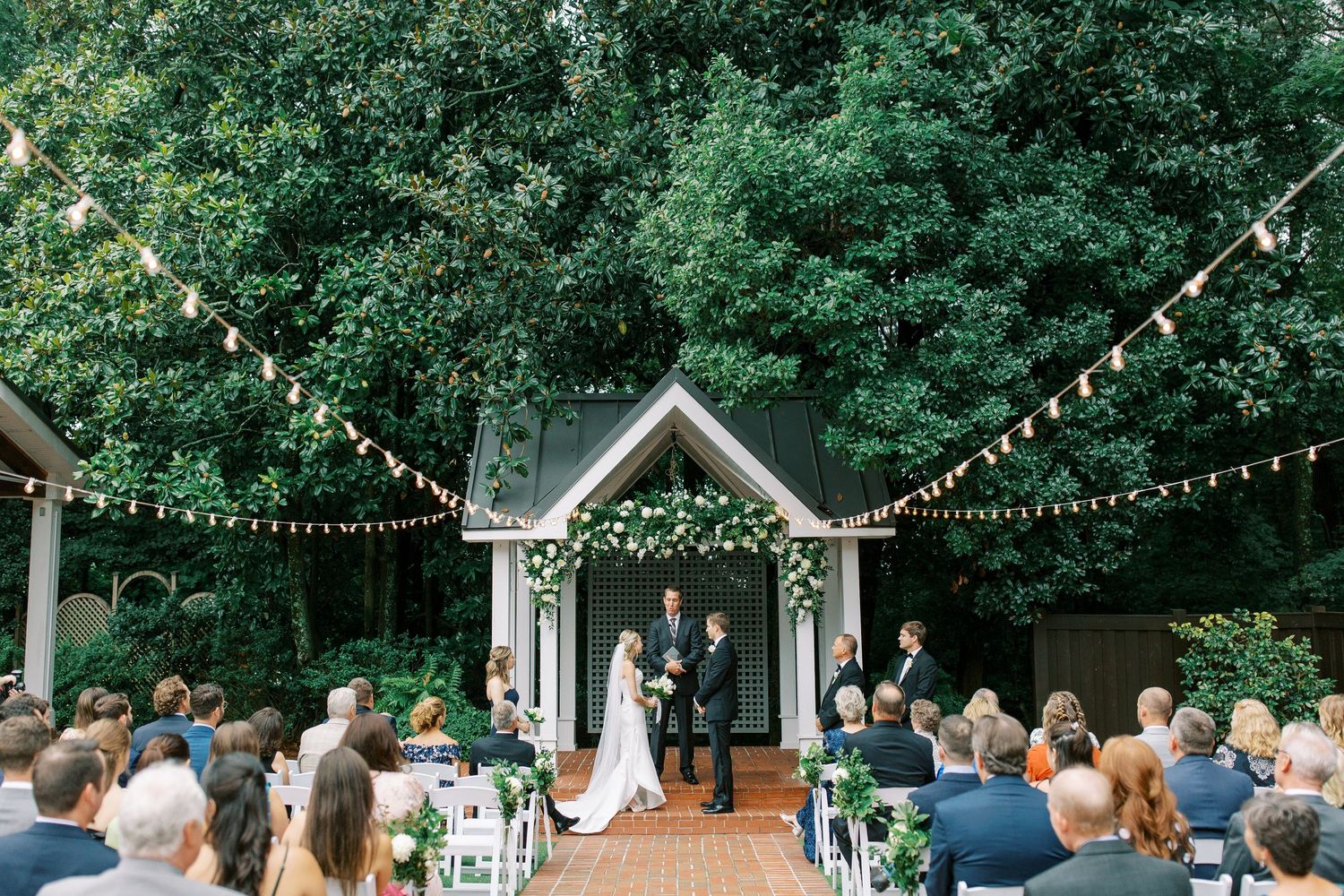 An outdoor wedding ceremony under string lights with guests seated in white chairs facing a wooden arbor.