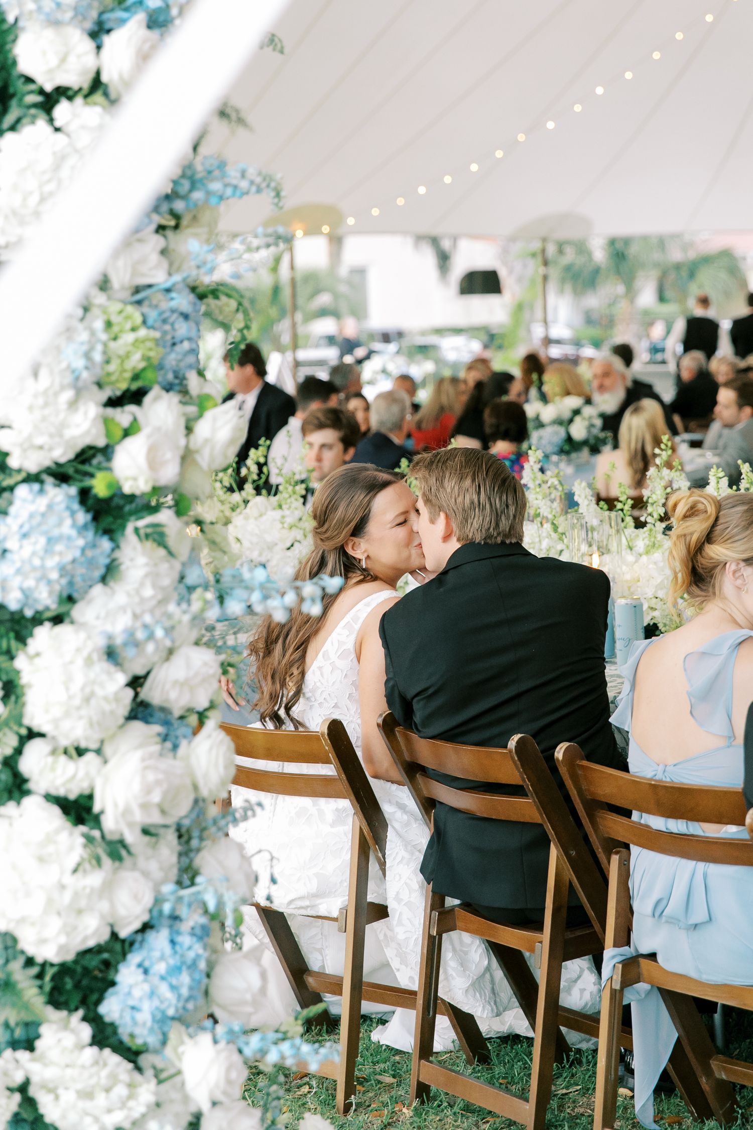 Romantic wedding reception under white tent with blue and white floral arrangements and string lights.