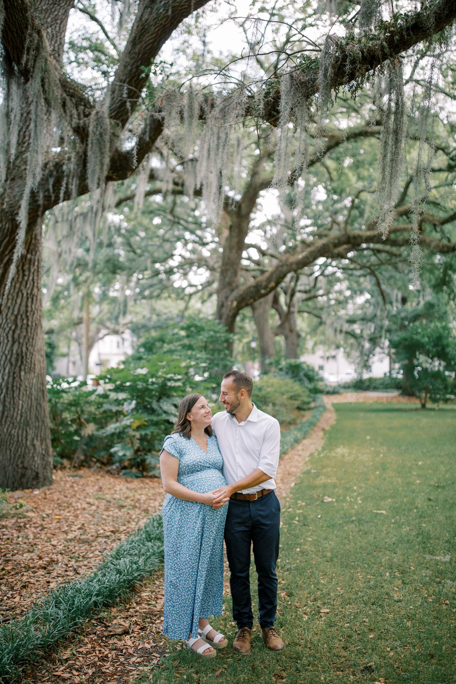 An expectant couple shares a romantic moment under moss-draped trees in a park setting.