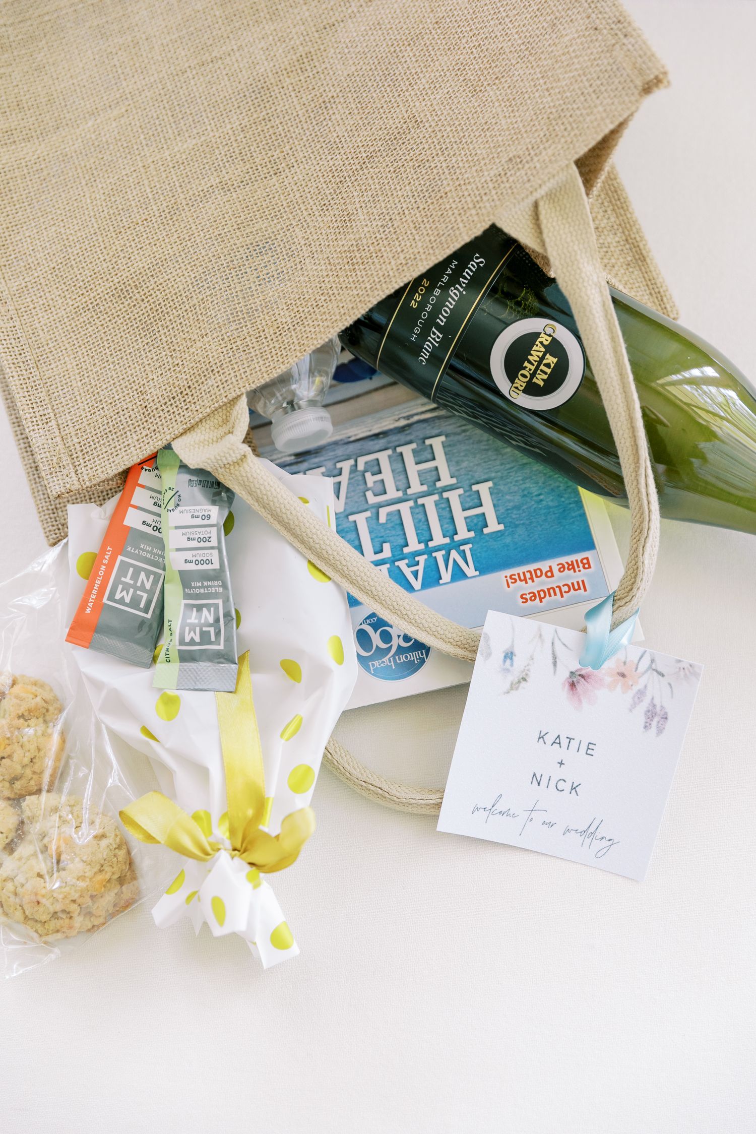 Welcome gift bag contents including a book, snacks, and card spilling out of a natural burlap tote on white background.