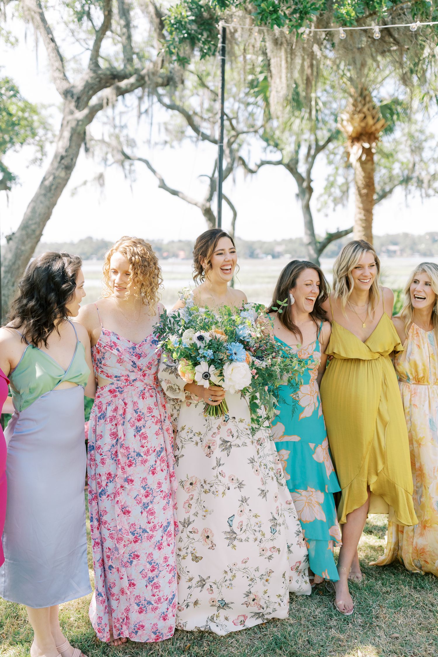 A group in colorful floral and pastel dresses pose together outdoors under mossy oak trees.