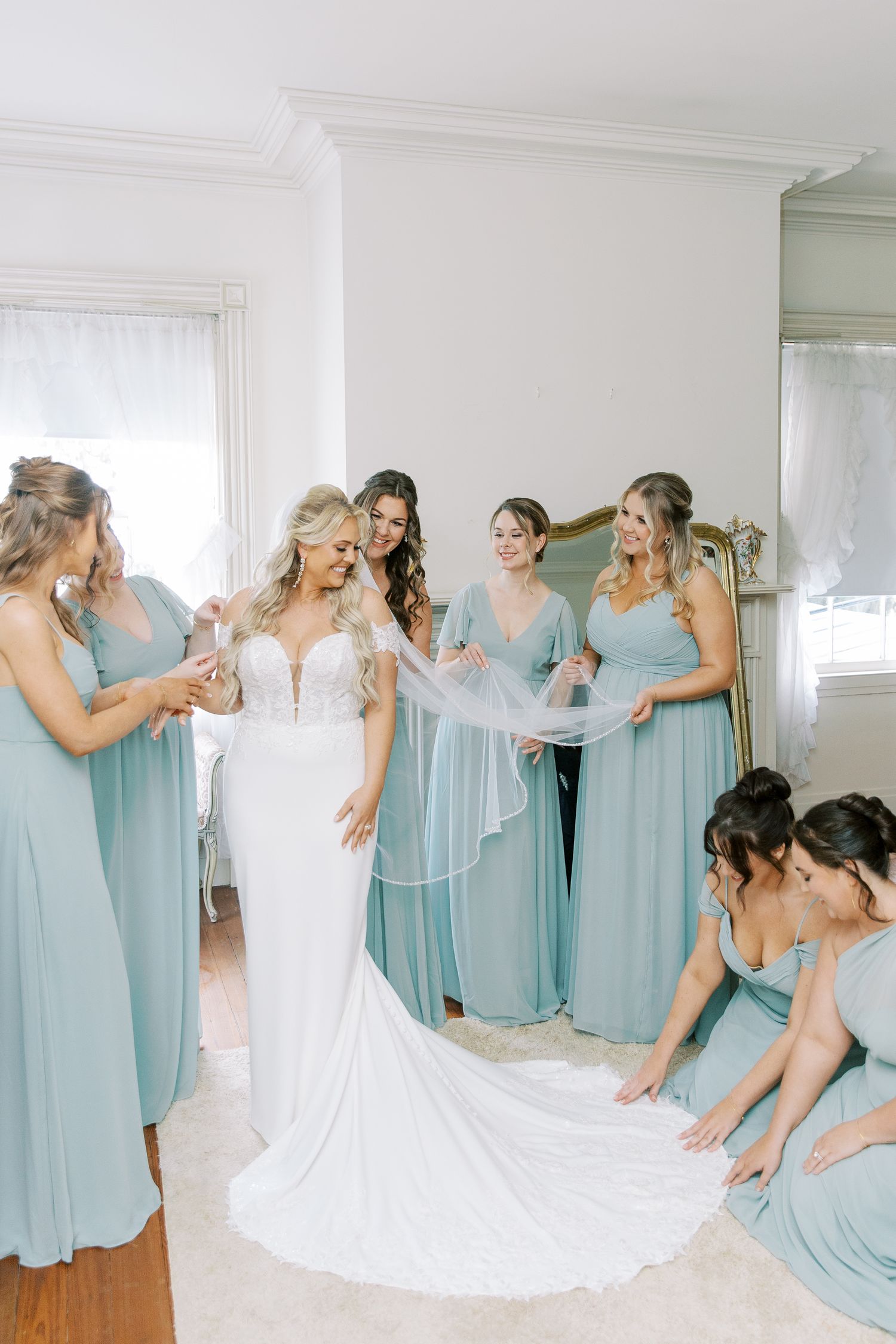 Wedding party in light blue bridesmaid dresses helping with a white lace wedding gown.
