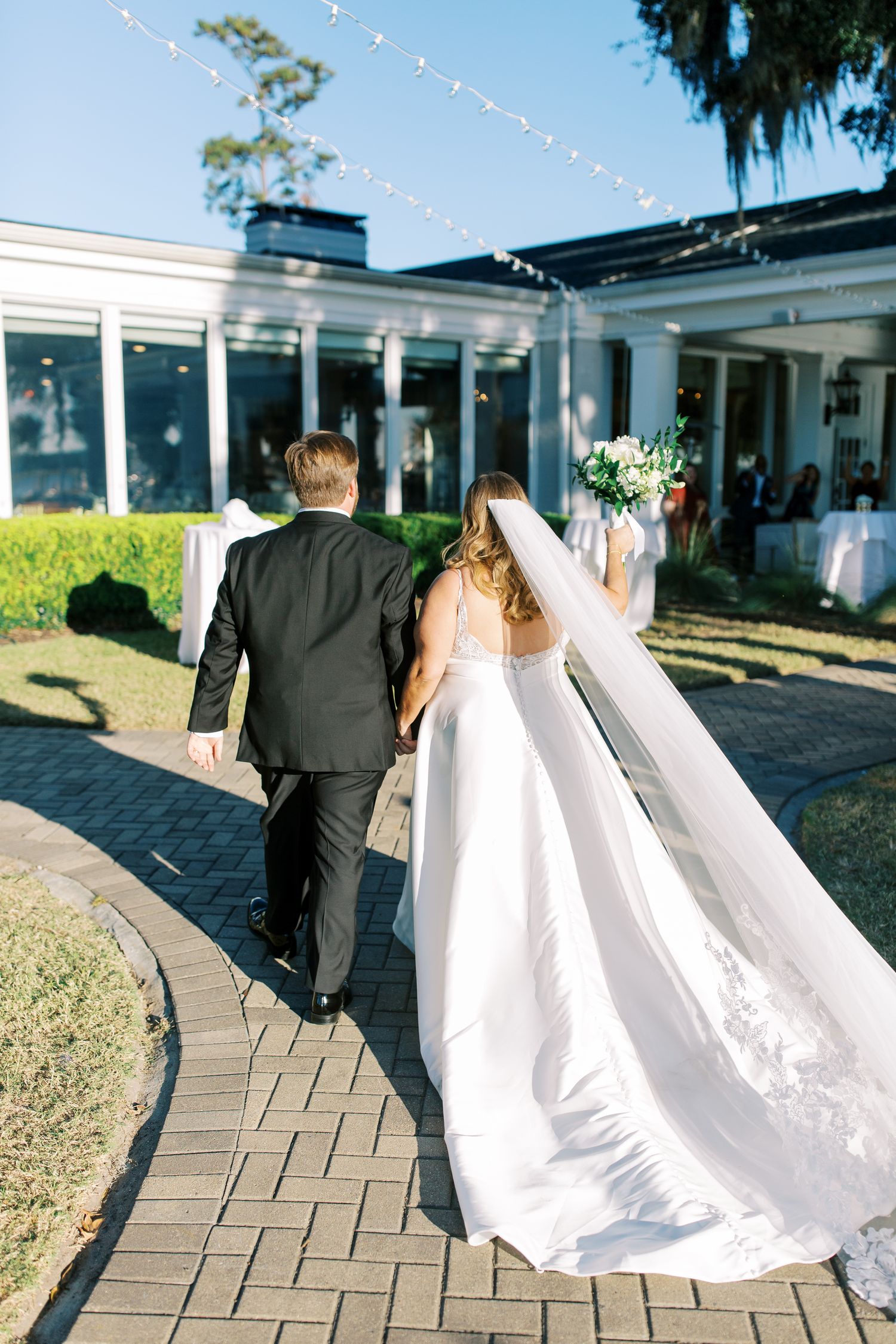 Newlywed couple walking down a curved brick path outside an elegant event venue with string lights.